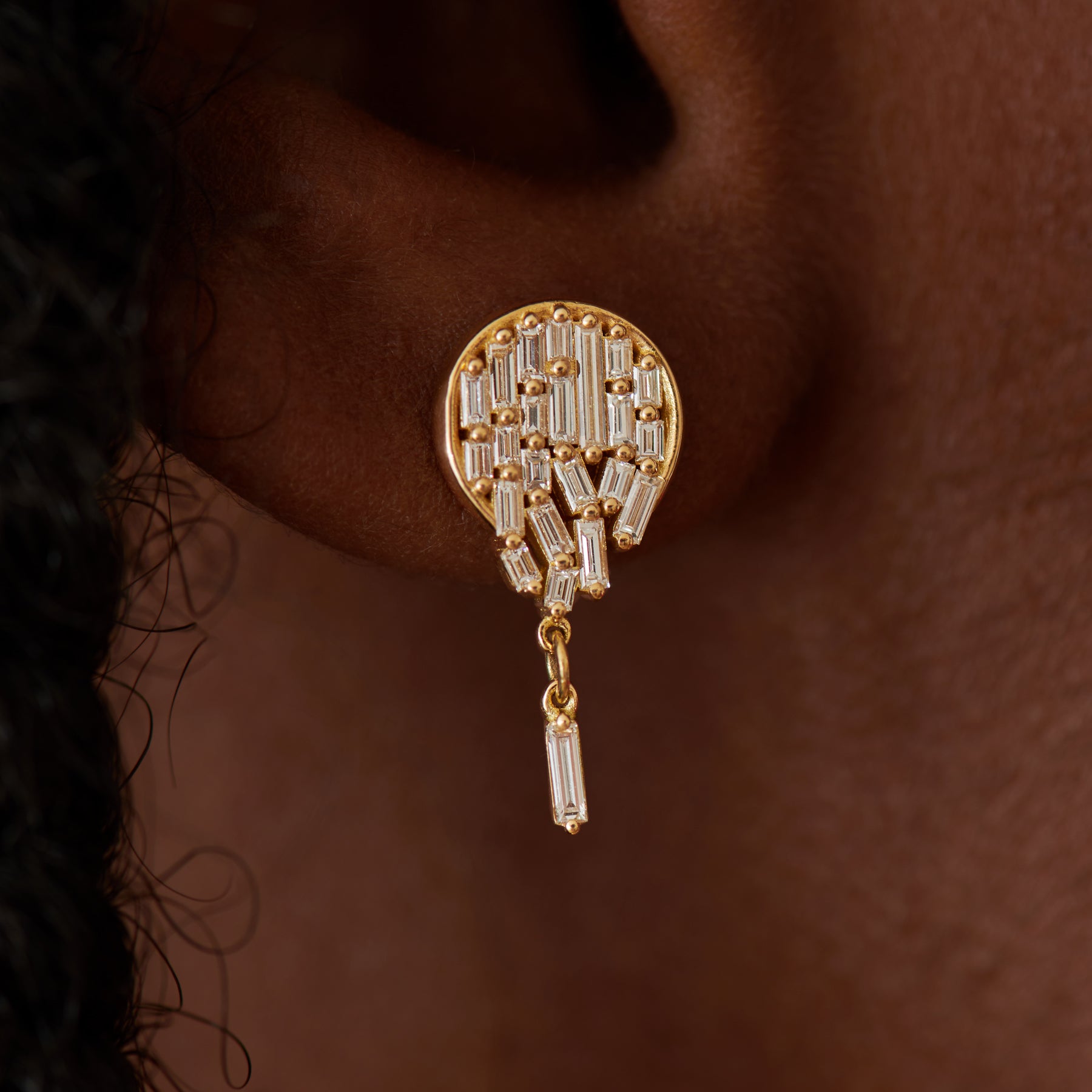 Gold earring with diamond accents worn on an ear against a brown background