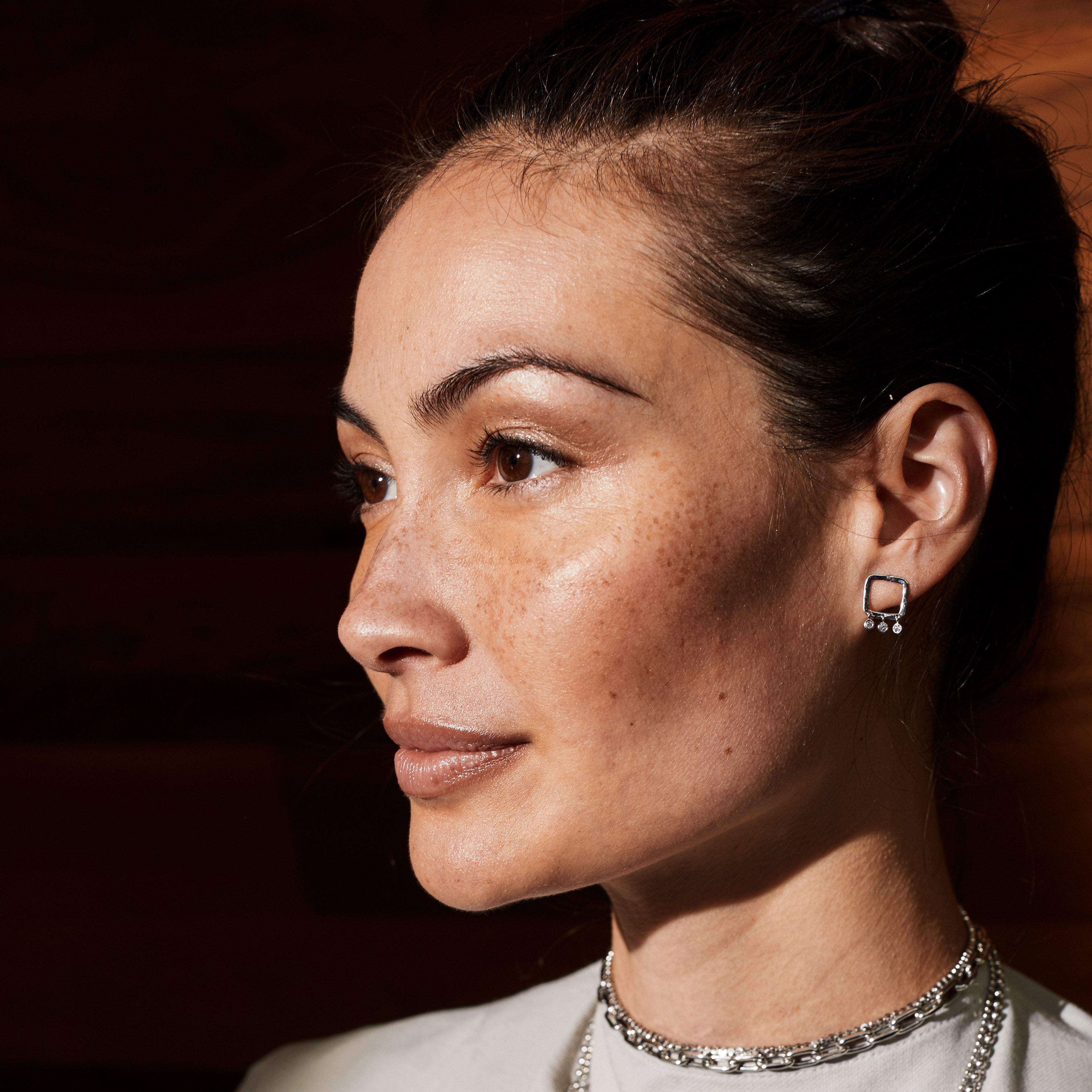 Woman wearing layered white gold necklaces and open square post earrings with diamond dangles against a dark background