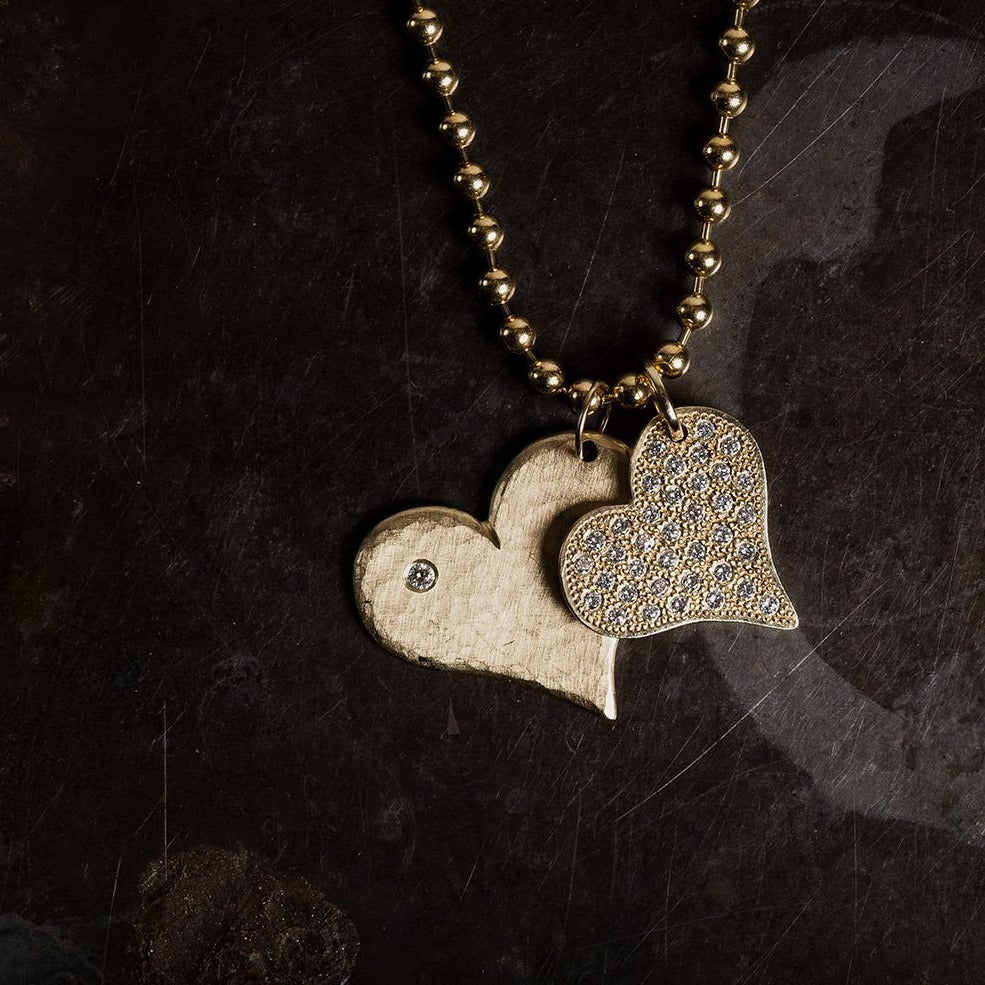 14k yellow gold hammered heart-shaped pendant with a white diamond on a ball chain paired with heart shaped pendant with beaded texture and paved set white diamonds on natural dark background