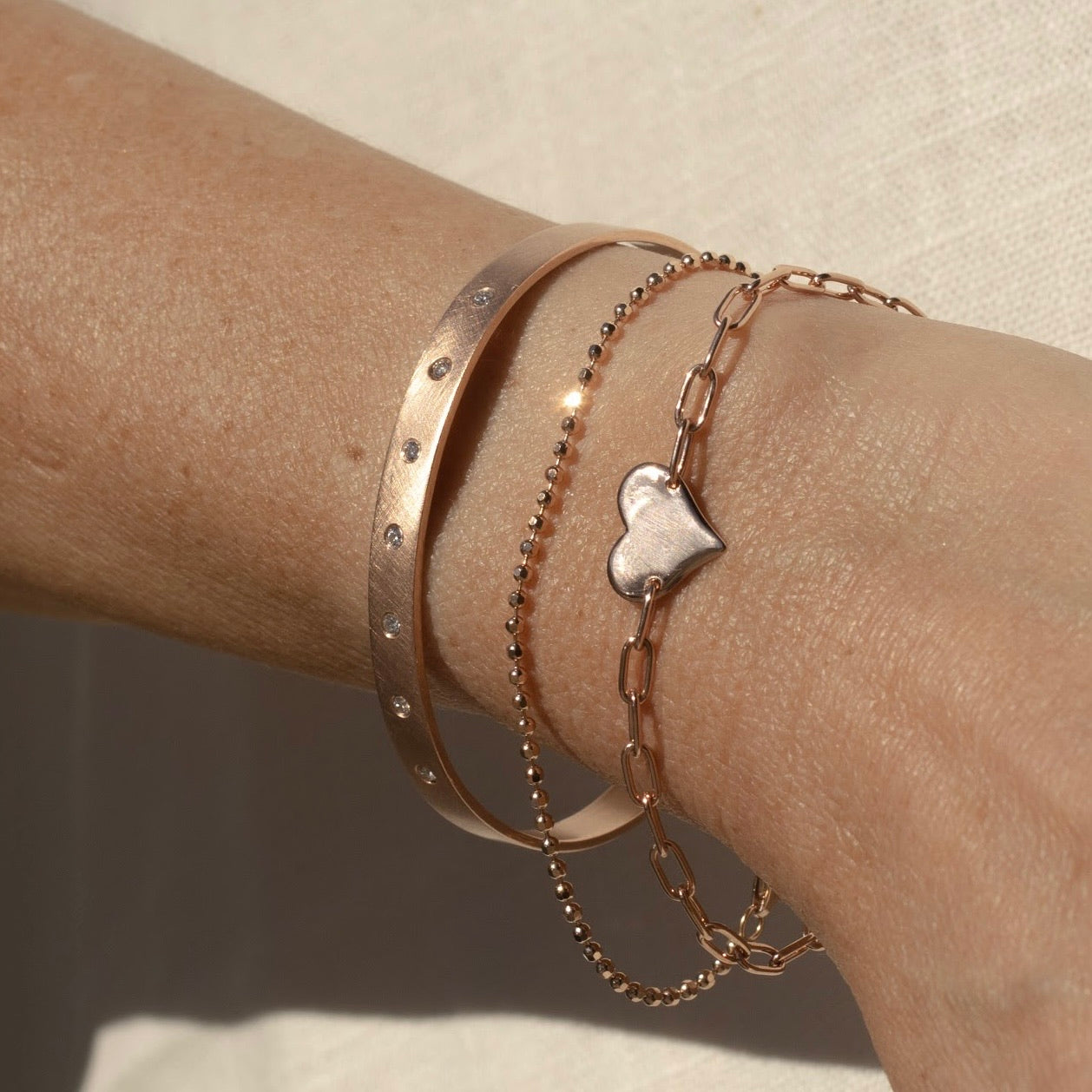14k rose gold heart bracelet with paperclip link chain stacked with diamond cut ball chain bracelet and satin cuff with white diamonds on model in lifestyle shot