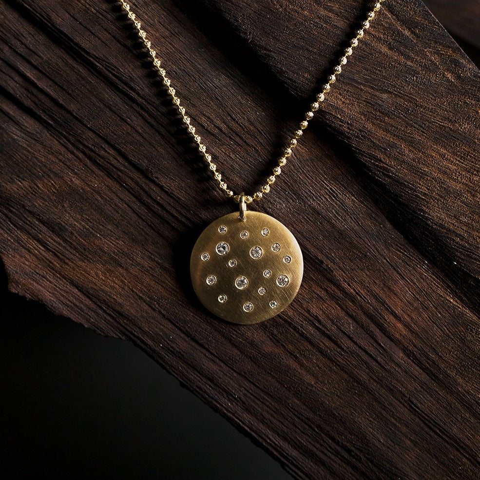14k yellow gold round pendant featuring scattered white diamonds on moca chain on natural wood background
