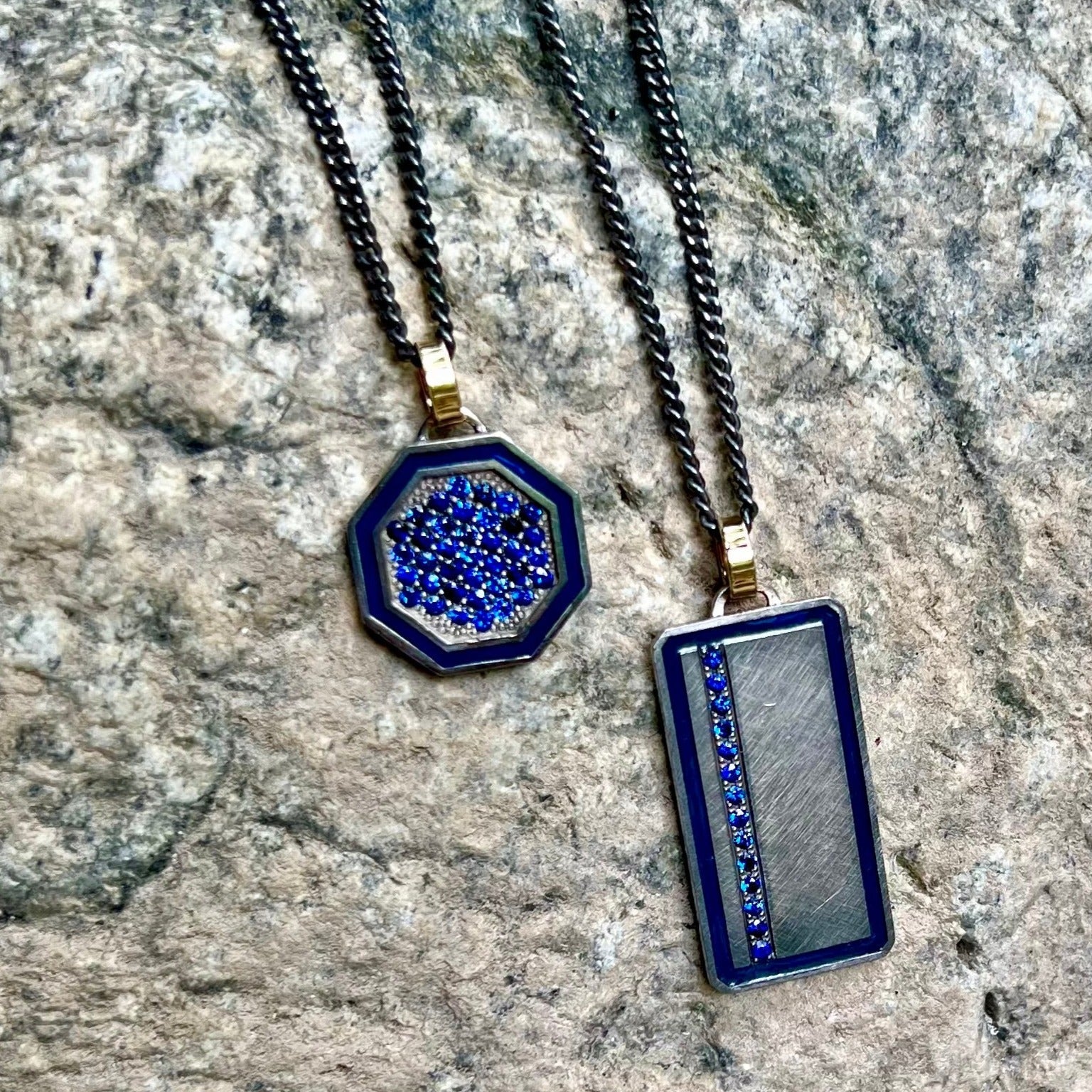 blackened sterling silver octagon pendant with colored gemstones and colored enamel border on 14k yellow gold jump ring besides blackened sterling silver rectangle pendant with colored enamel border and blue gemstones with 14k yellow gold jump ring on stone background