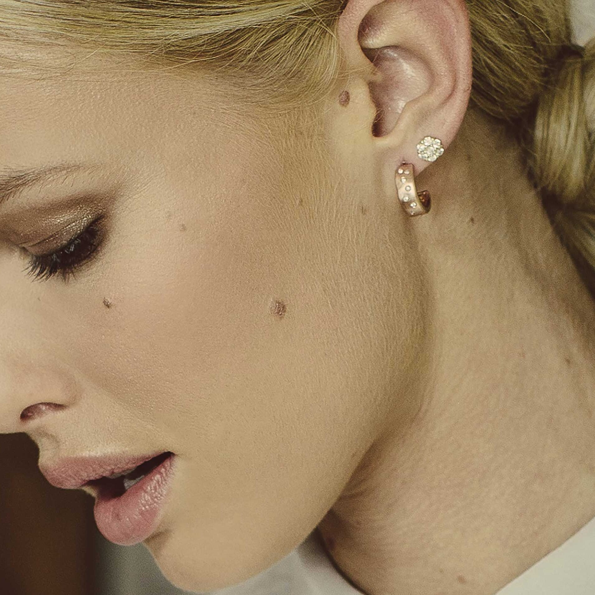 Close-up of a model wearing 14k gold huggie hoops with scattered diamonds and cluster earrings with a blurred background