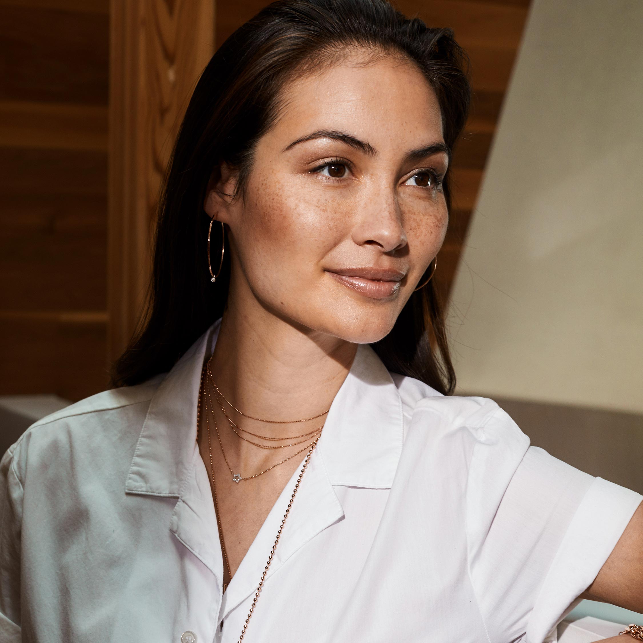Lifestyle shot of woman wearing a white shirt and layered  14k yellow gold diamond jewelry by Julez Bryant with a neutral background