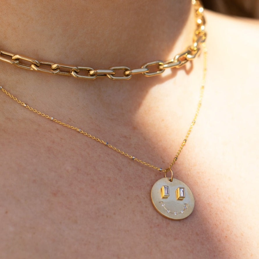 14k yellow gold smiley face pendant with baguette diamonds stacked with mica chain on model in lifestyle shot