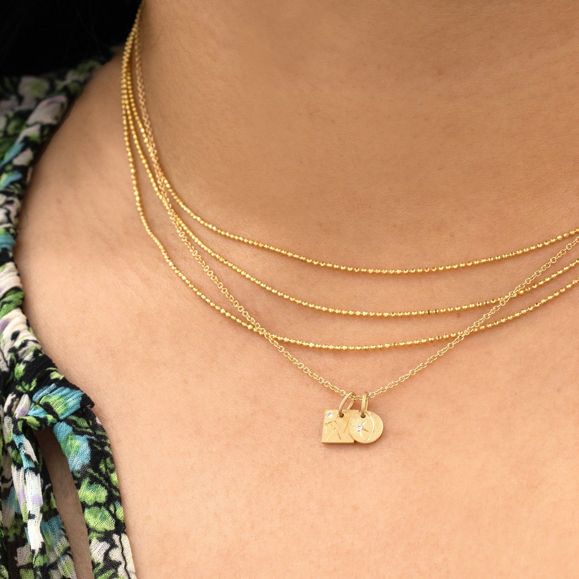 14k yellow gold square pendant with hand etched mountains and sun with white diamond paired with round pendant charm stacked with the chai necklace on model in lifestyle shot