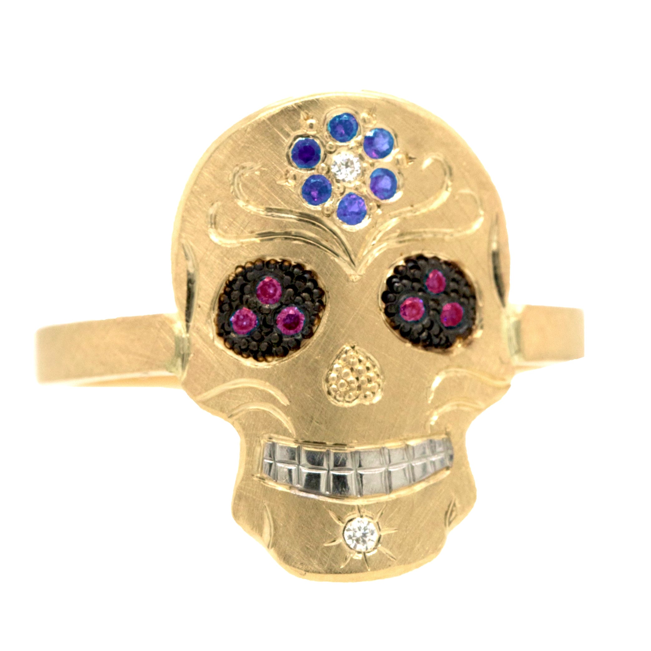 Gold skull-shaped ring with gemstones on a white background. Pave set bluesapphire flower on forehead, pave set eyes and diamond on chin. Fine engraving and milgrain detail.