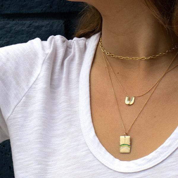 Close-up of a person wearing gold necklaces with green sapphire and diamond accents, arched rainbow designs unique pendants layered dark blue background