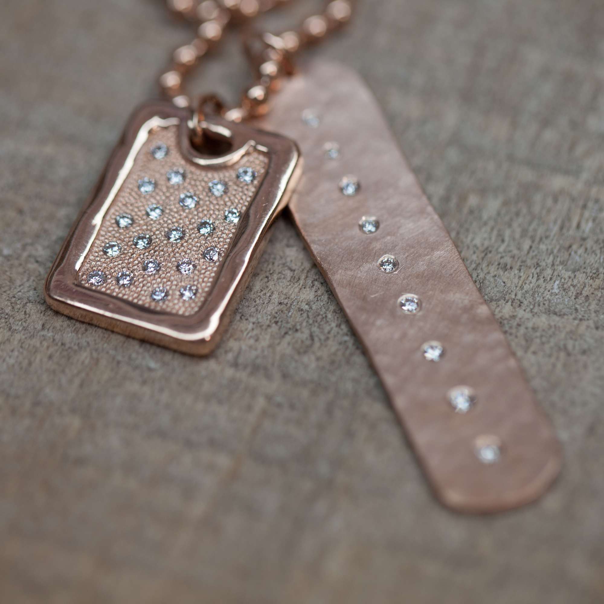 14k rose gold rectangular dog tag with shiny edges and milgrain texture in center with scattered white diamonds beside an elongated hammered oval pendant with a line of white diamonds down the center on tan background