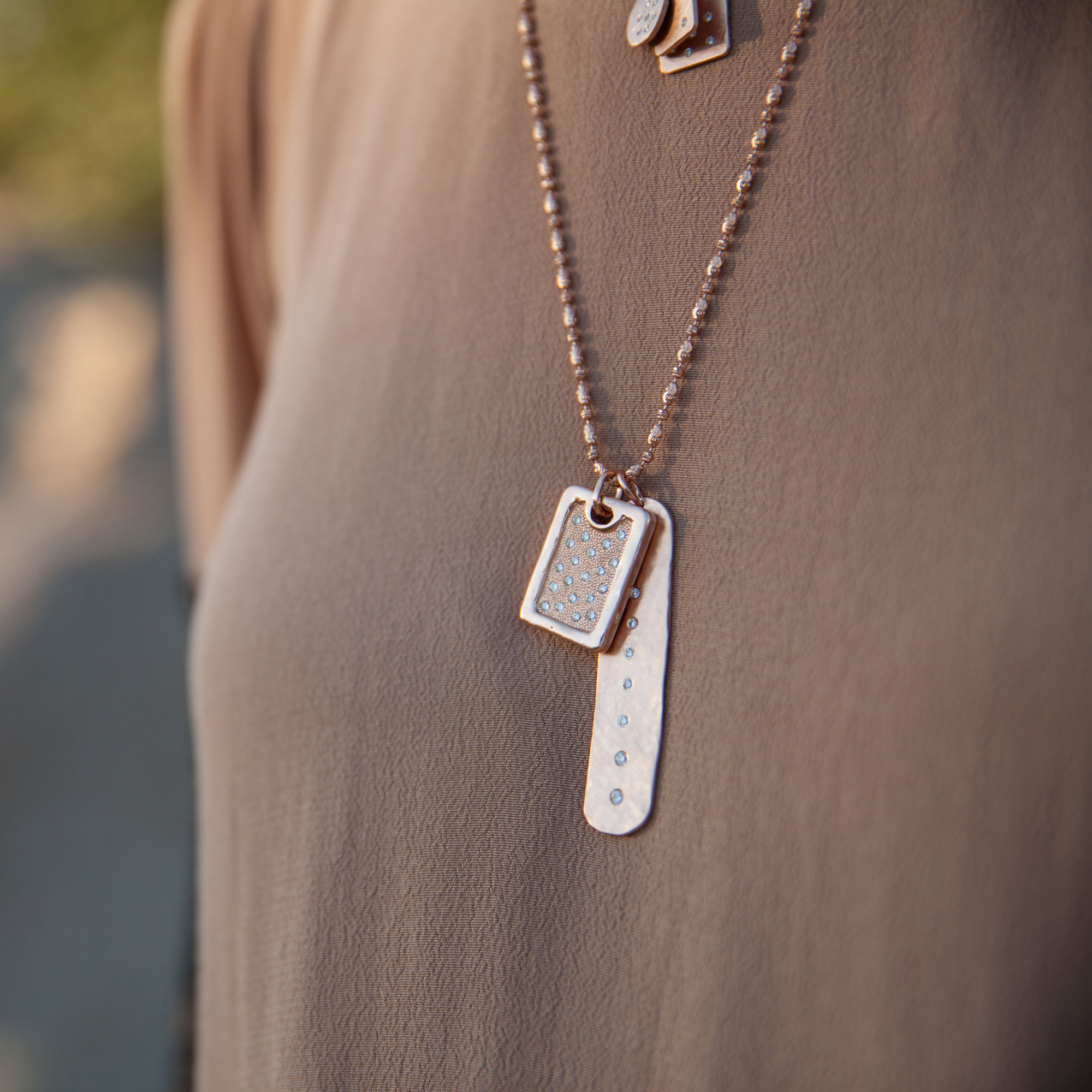 14k rose gold rectangular dog tag with shiny edges and milgrain texture in center with scattered white diamonds beside an elongated hammered oval pendant with a line of white diamonds down the center on model in lifestyle shot