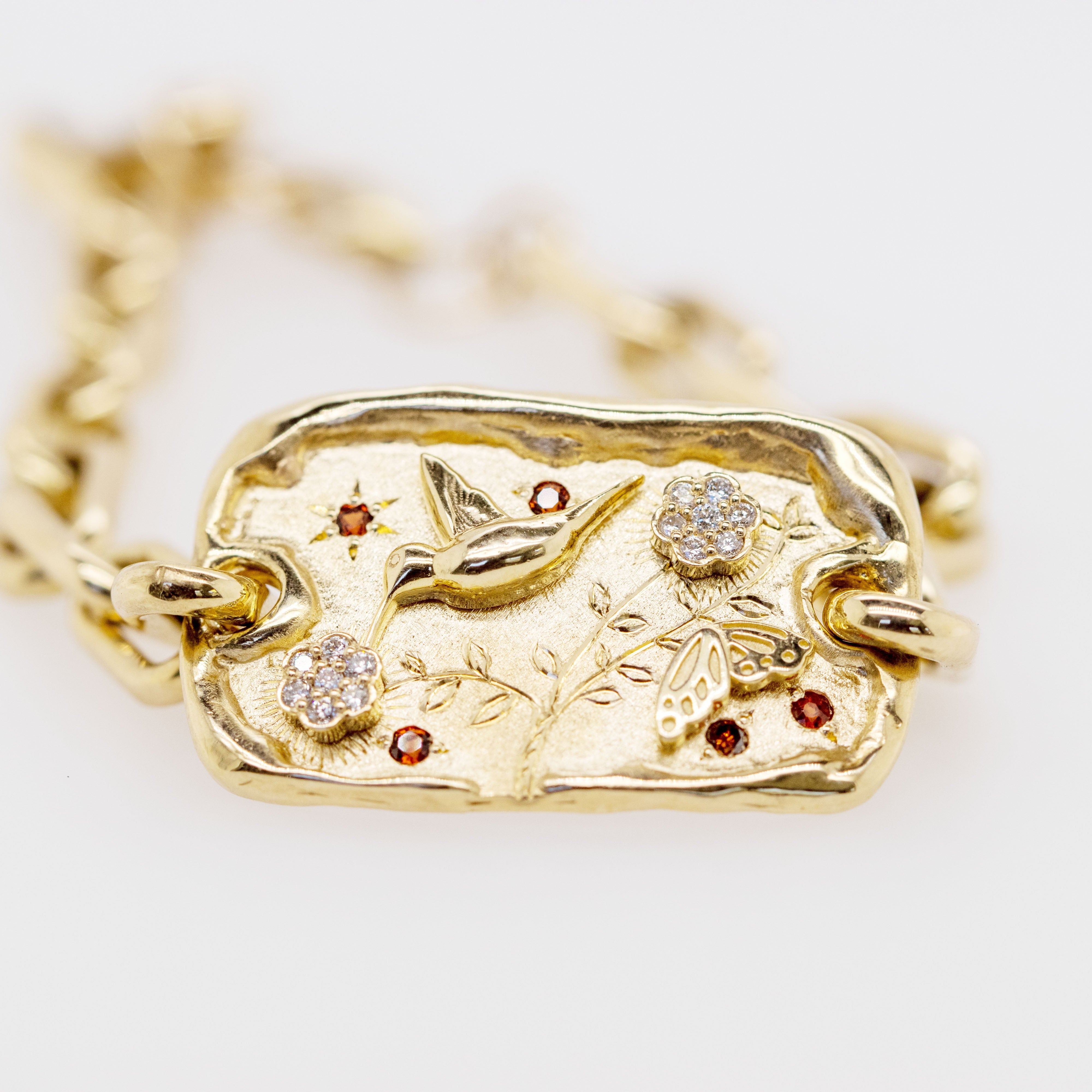 A gold chain bracelet featuring a textured rectangular pendant with a bird and floral motif, accented by small red gemstones and clear stones, shown on a white background.
