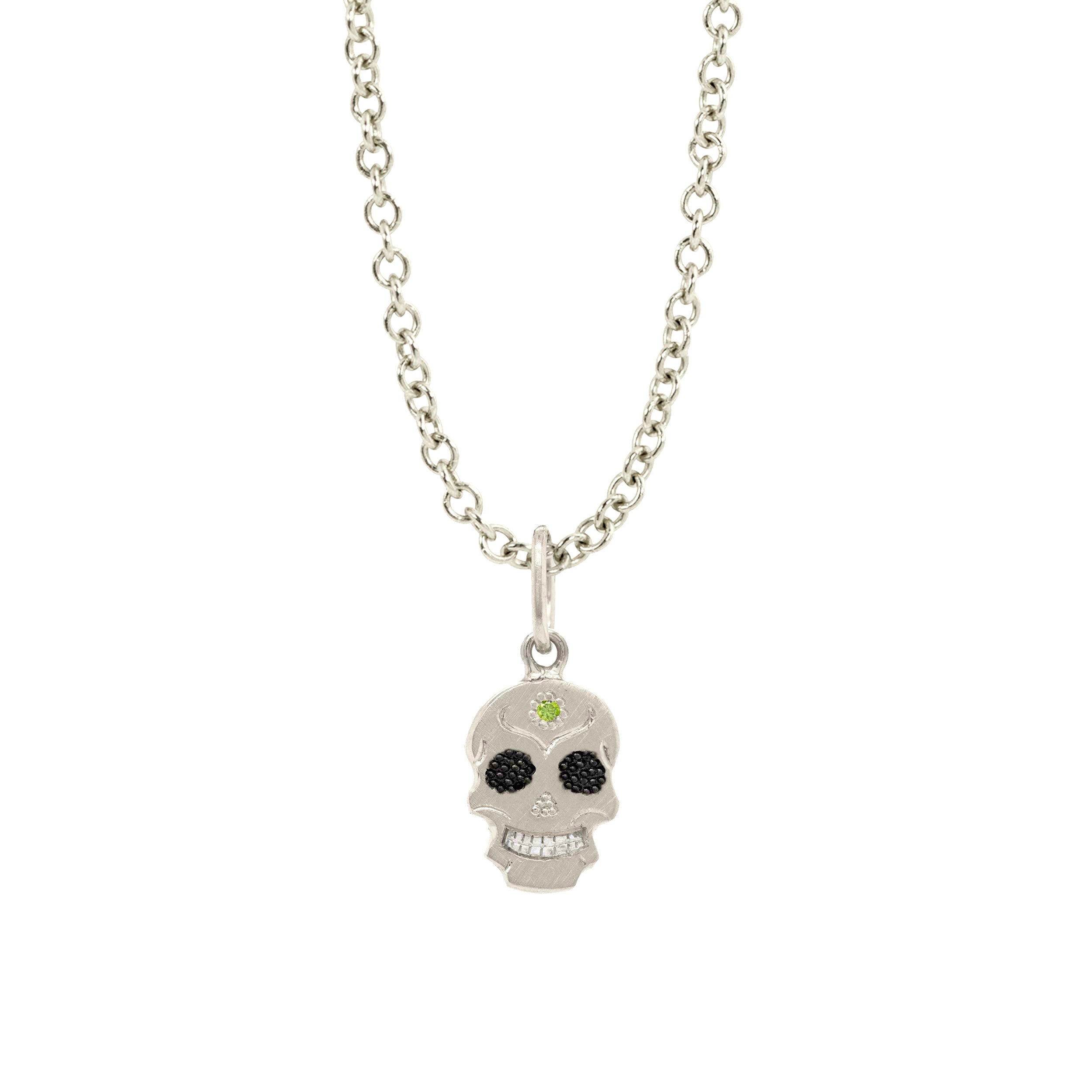 A Teeny Tiny collection white gold charm in the shape of a sugar skull with a round apple green diamond in the center, displayed on a gold chain against a white background.