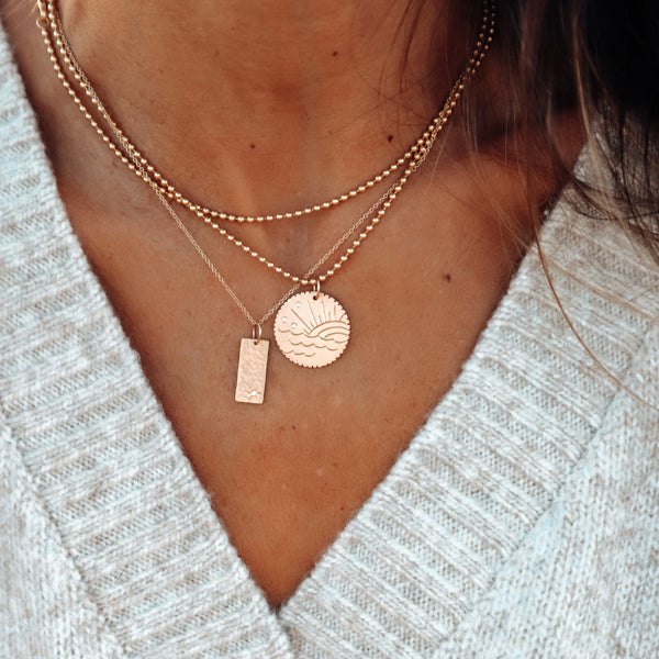 14k gold rectangular pendant with white diamond hand etched starburst on rolo chain paired with gone round charm with beach sunset engraving and white diamonds on ball chain in lifestyle shot on model