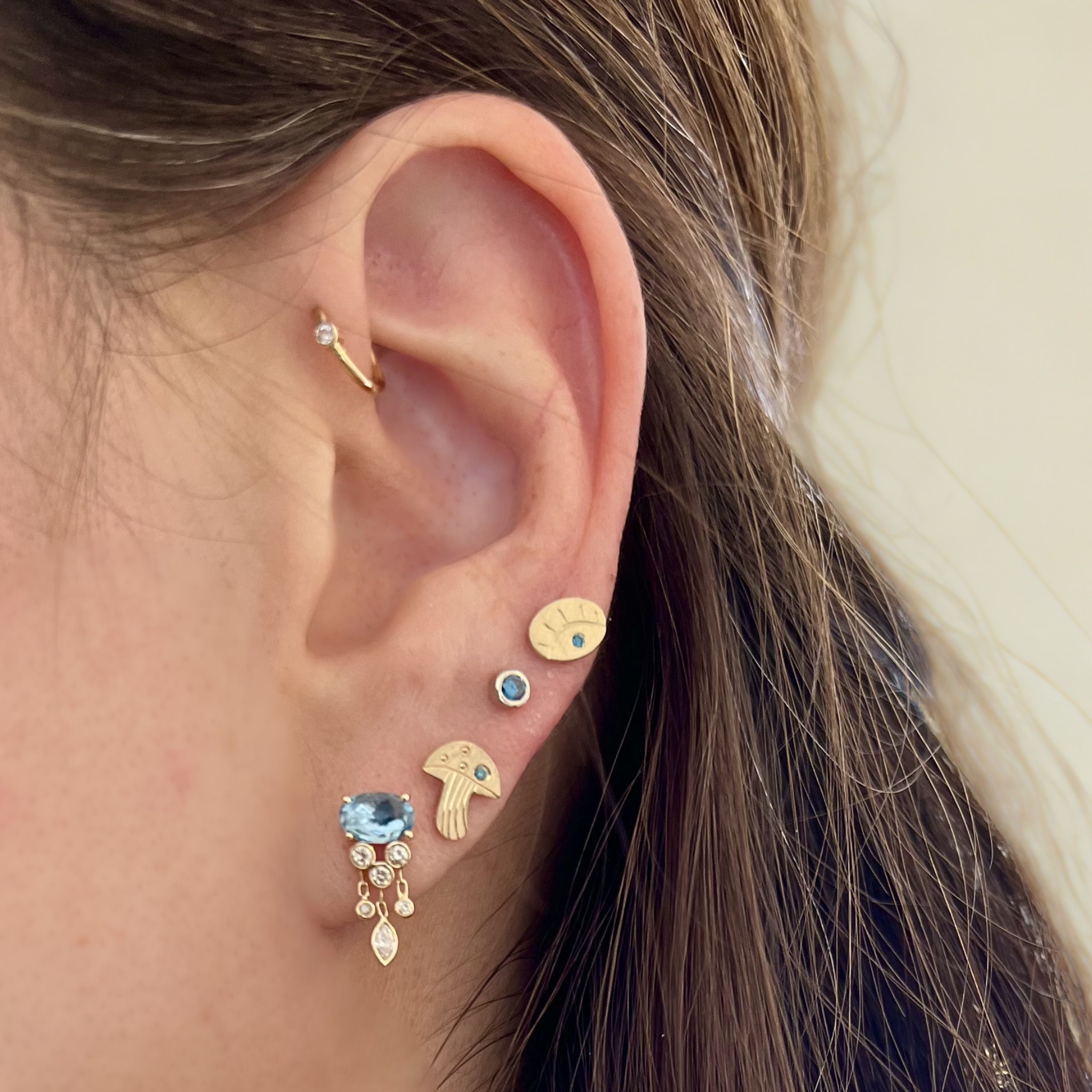 Close-up of an ear with multiple gold stud earrings with blue gemstones whimsical cut out shapes, blurred background.