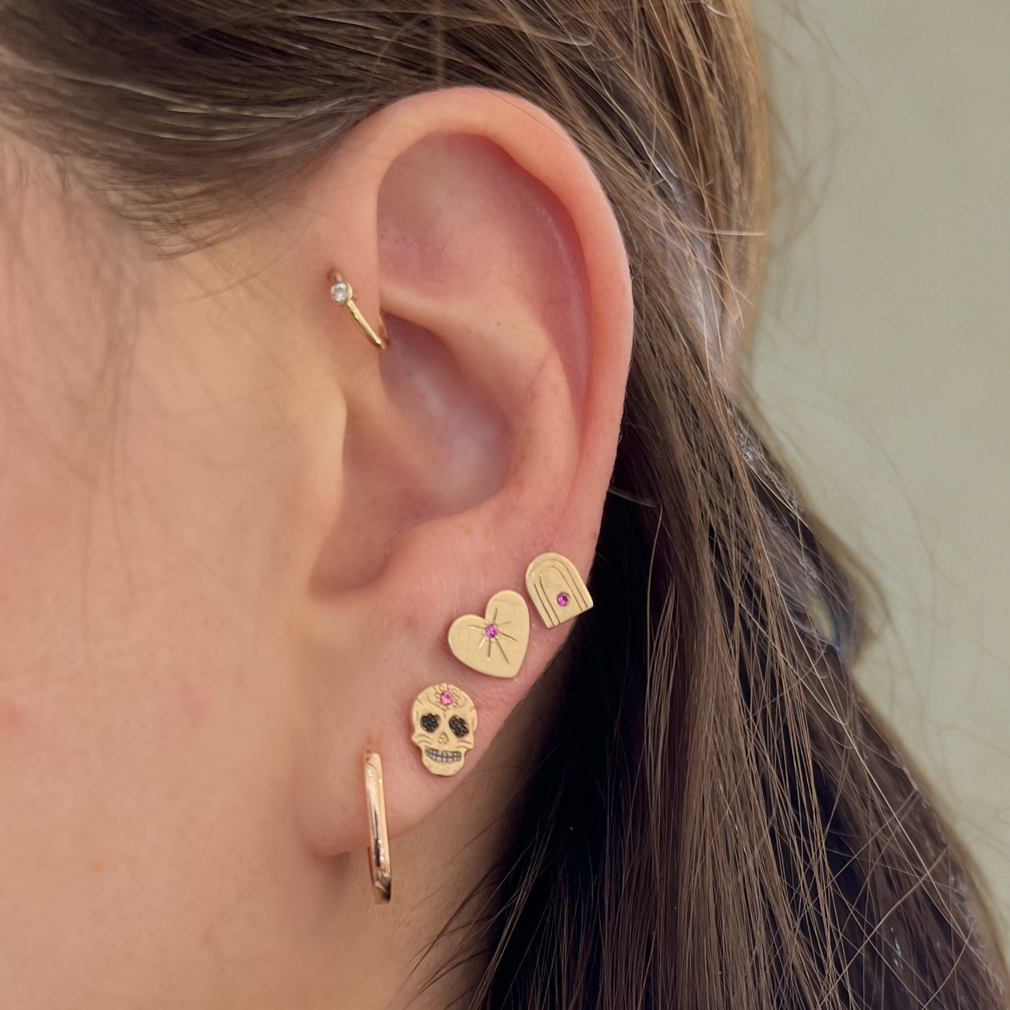 Close-up of an ear with gold earrings including a heart, skull, rainbow and hoop design with diamond and pink sapphire accents blurred background