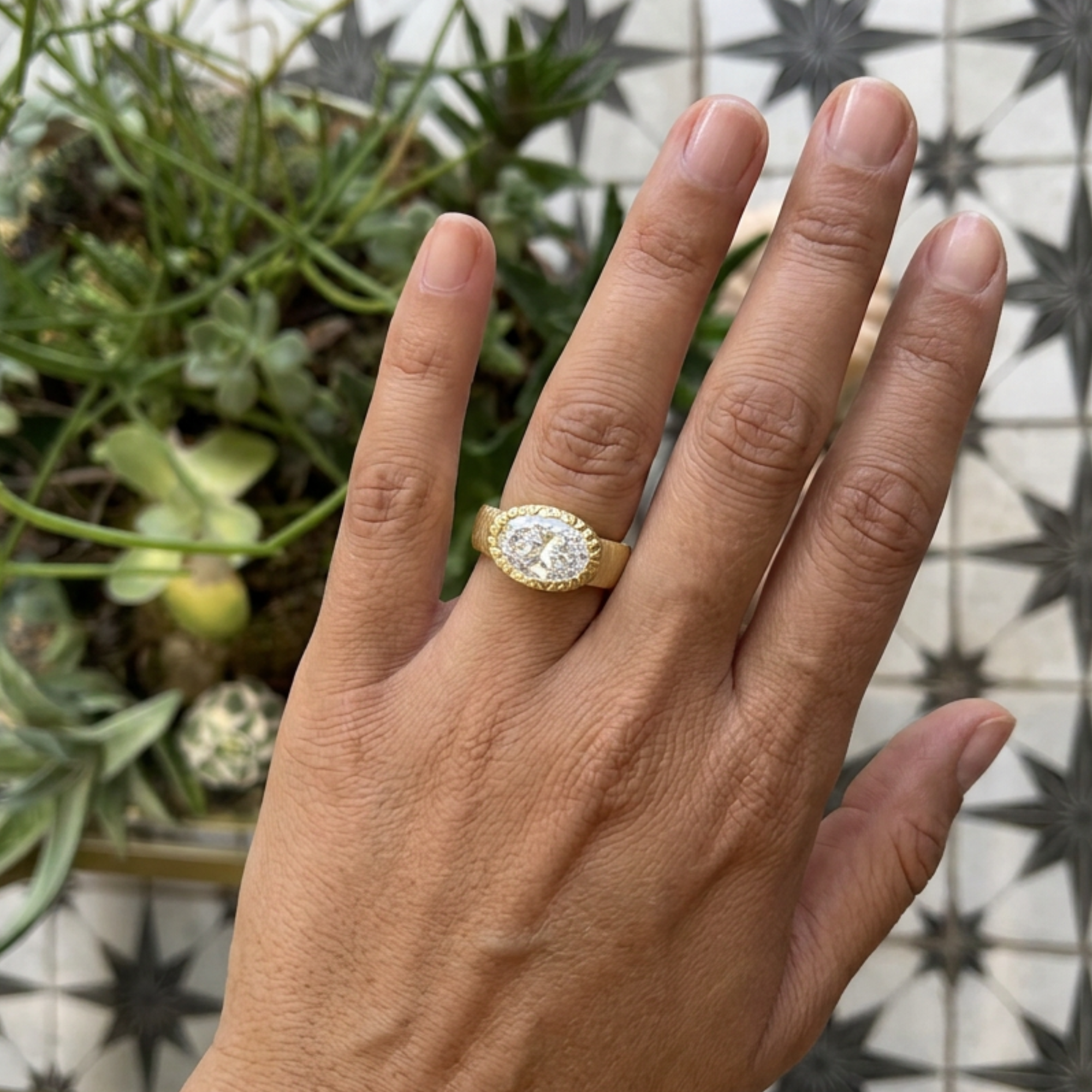 Image of a lab grown chunky east west oval bezel solitaire carved bezel engagement ring in 14k yellow gold on lifestyle shot with blurred greenery worn on a model.