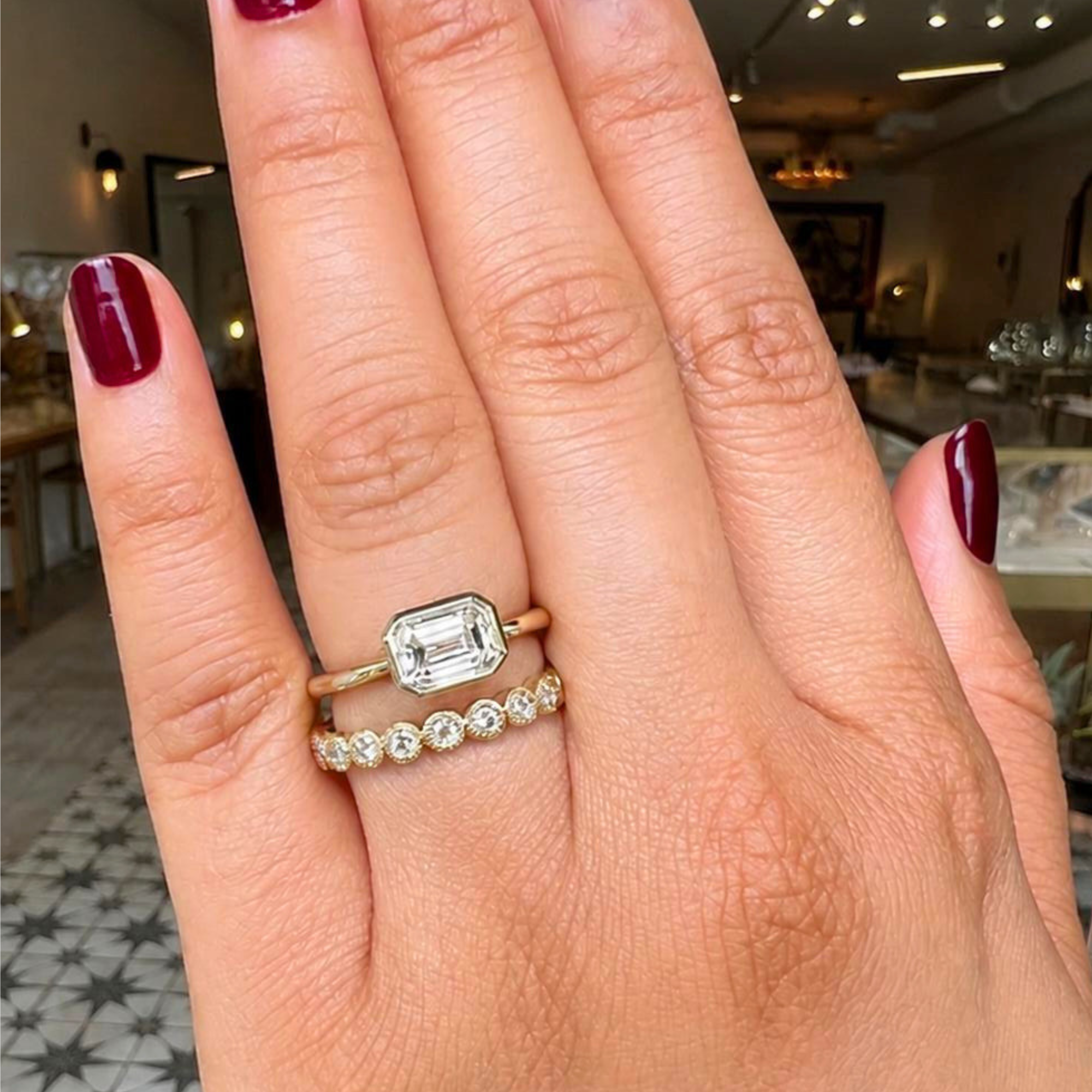 Image of a lab grown emerald cut cut bezel solitaire east west tapered 14k yellow gold engagement ring candid with blurred background.
