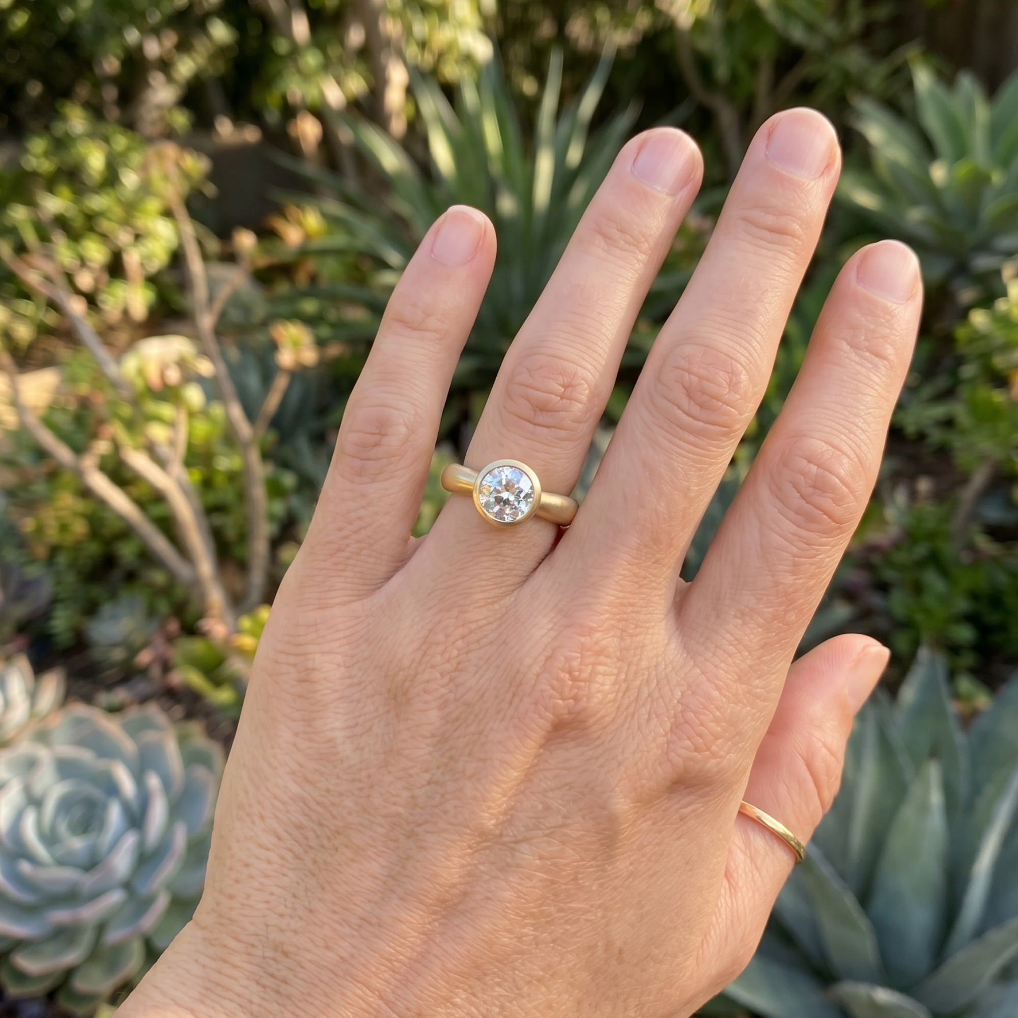 Image of a lab grown round brilliant bezel solitaire wide band14k yellow gold engagement ring candid with blurred greenery background.