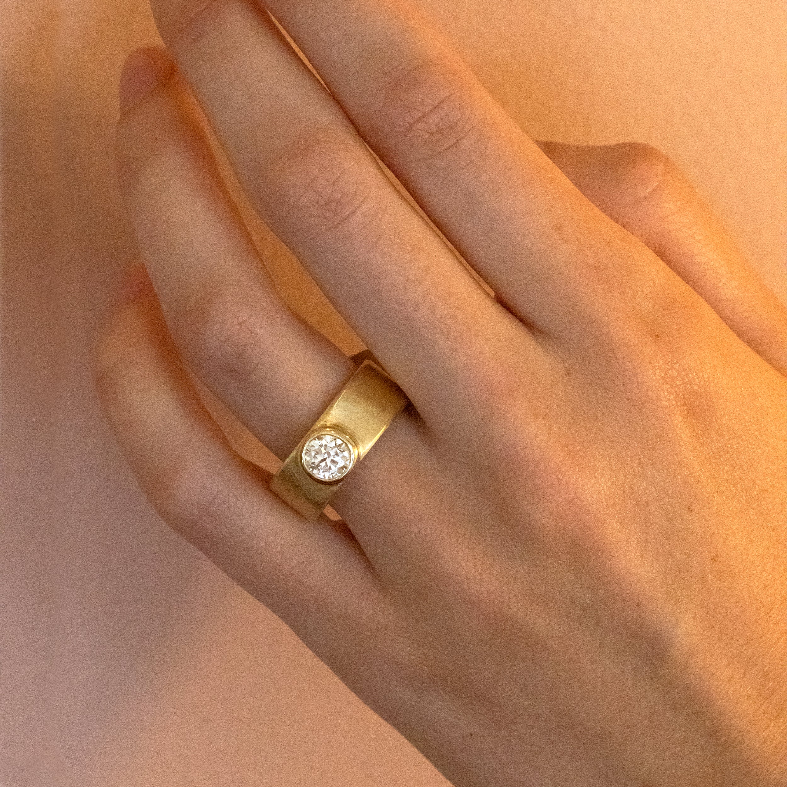 Wide cigar band bezel solitaire diamond engagement ring with satin finish close up on models hand blush background