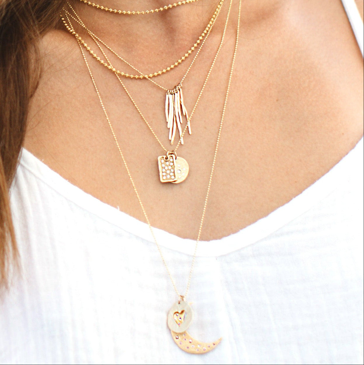 Close-up of a woman wearing multiple gold necklaces with heart and moon pendants dainty 1mm diamond ball and 2mm ball chain on a white background.