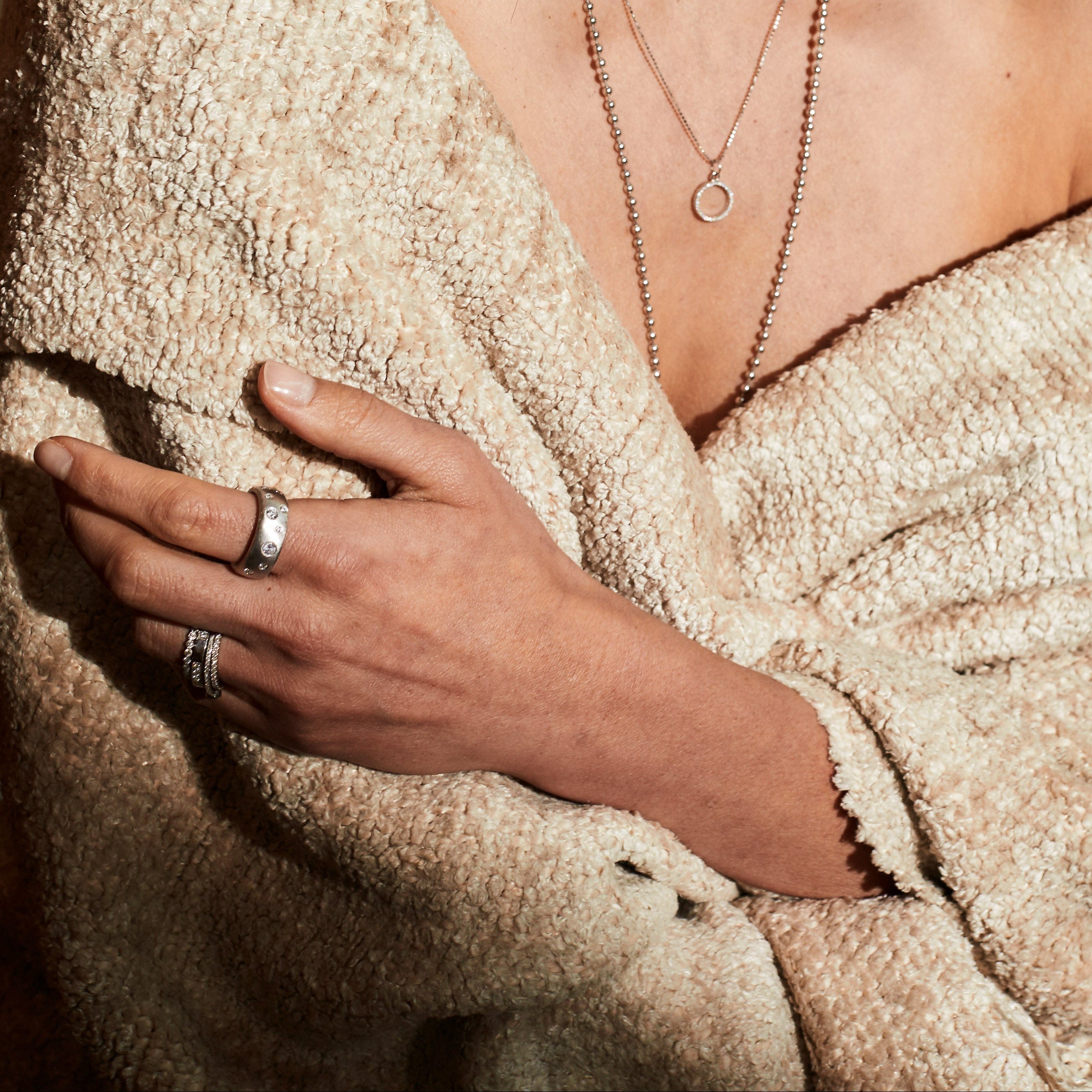 Woman wrapped in a beige blanket with layered white gold necklaces, rings with white diamonds blurred background
