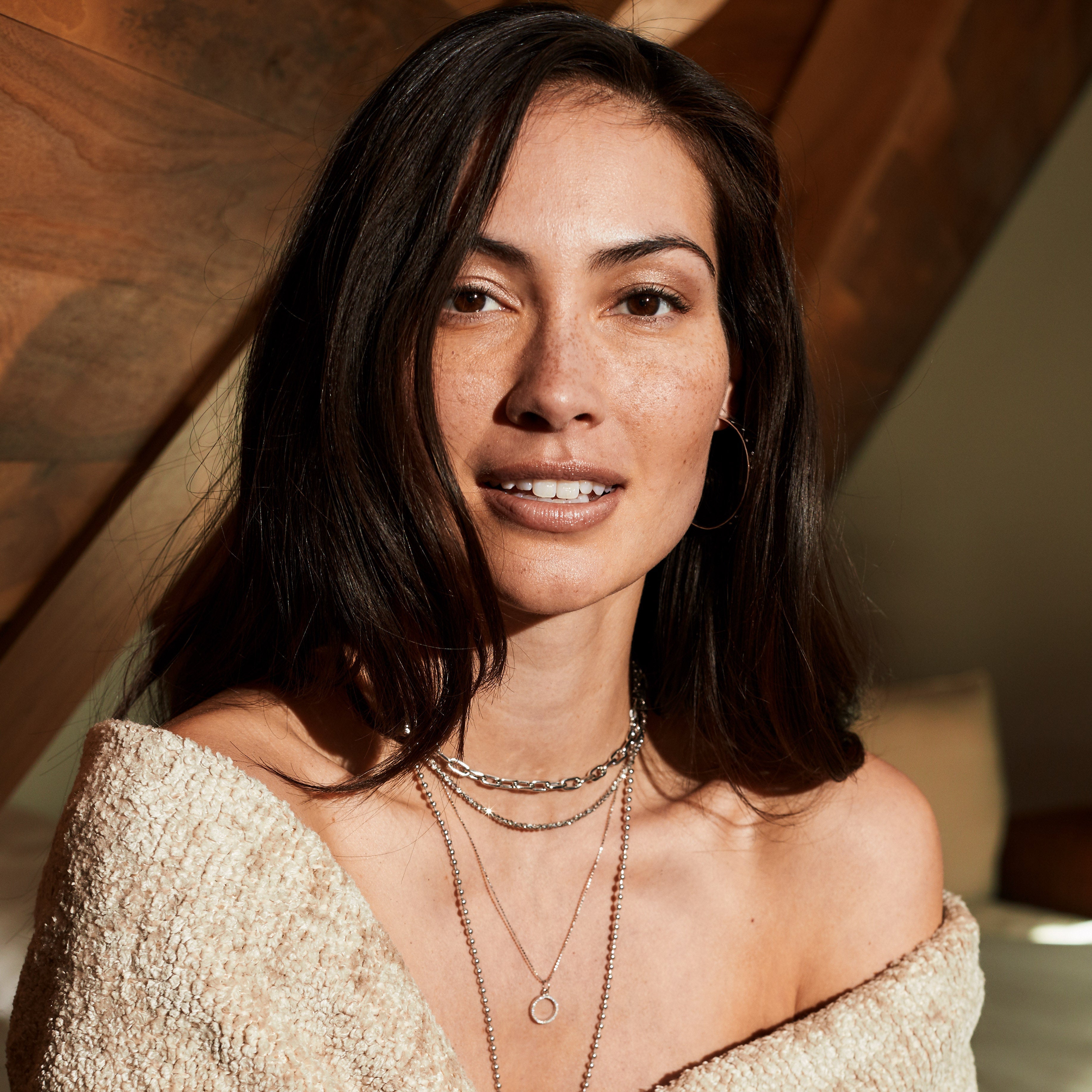 Woman wrapped in a beige blanket with layered necklaces, rings, hoops silver colored white gold with diamonds, indoors.