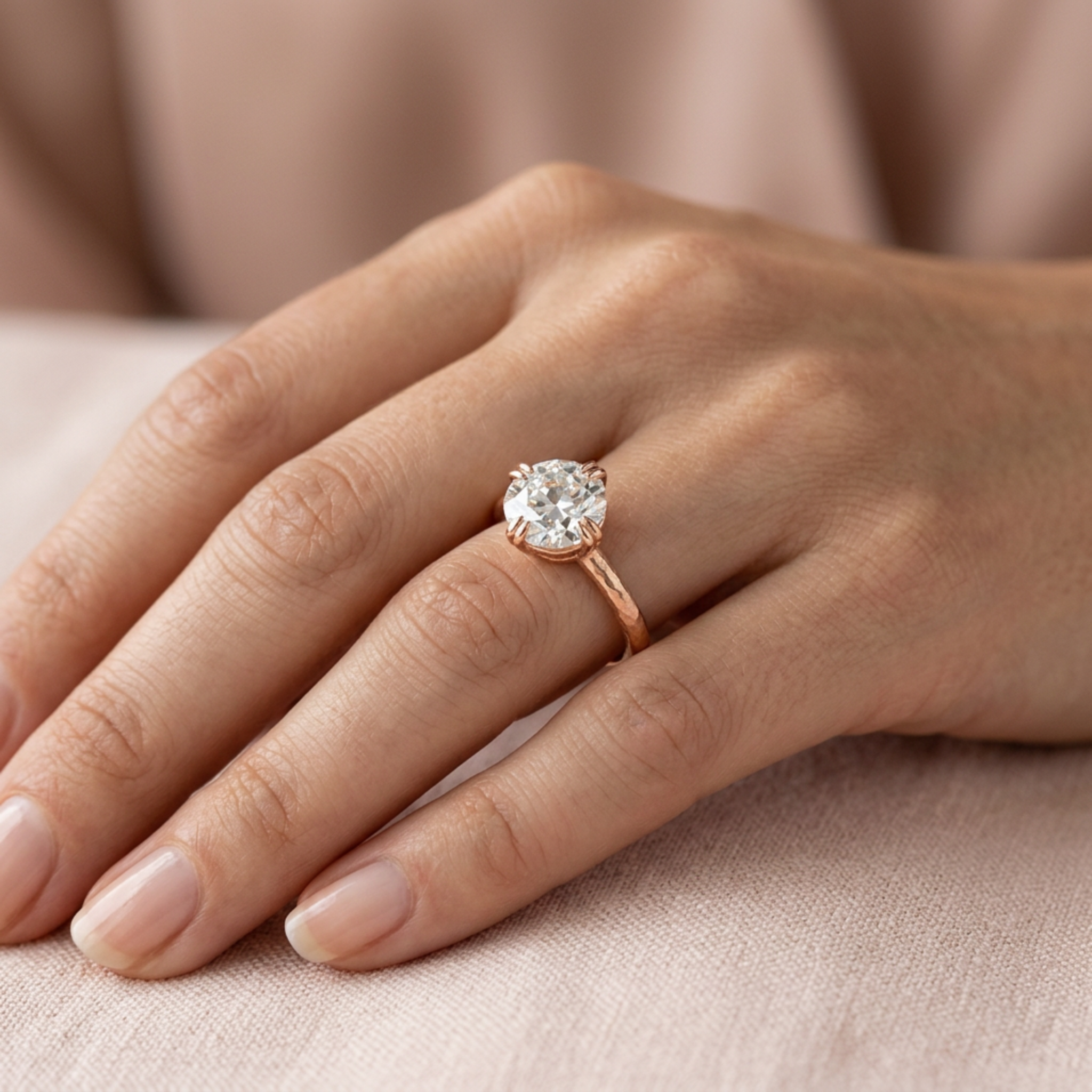 Close up of hand wearing a 14k gold solitaire engagement ring with a double claw prong setting featuring an old Euro cut white diamond on a thin band with a satin finish soft blush background