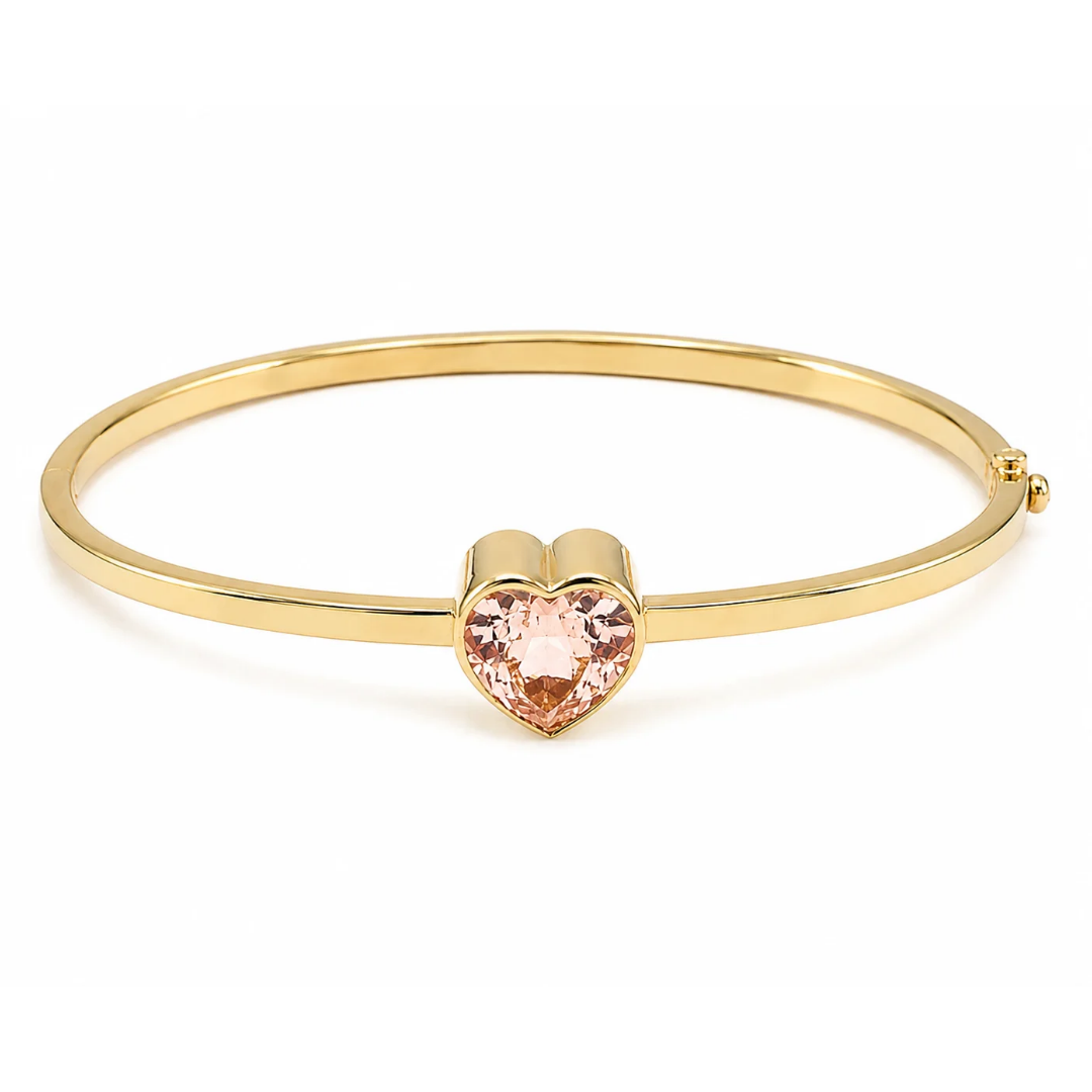 A gold-plated bangle bracelet featuring a polished heart-shaped pink gemstone in the center, set against a clean white background.