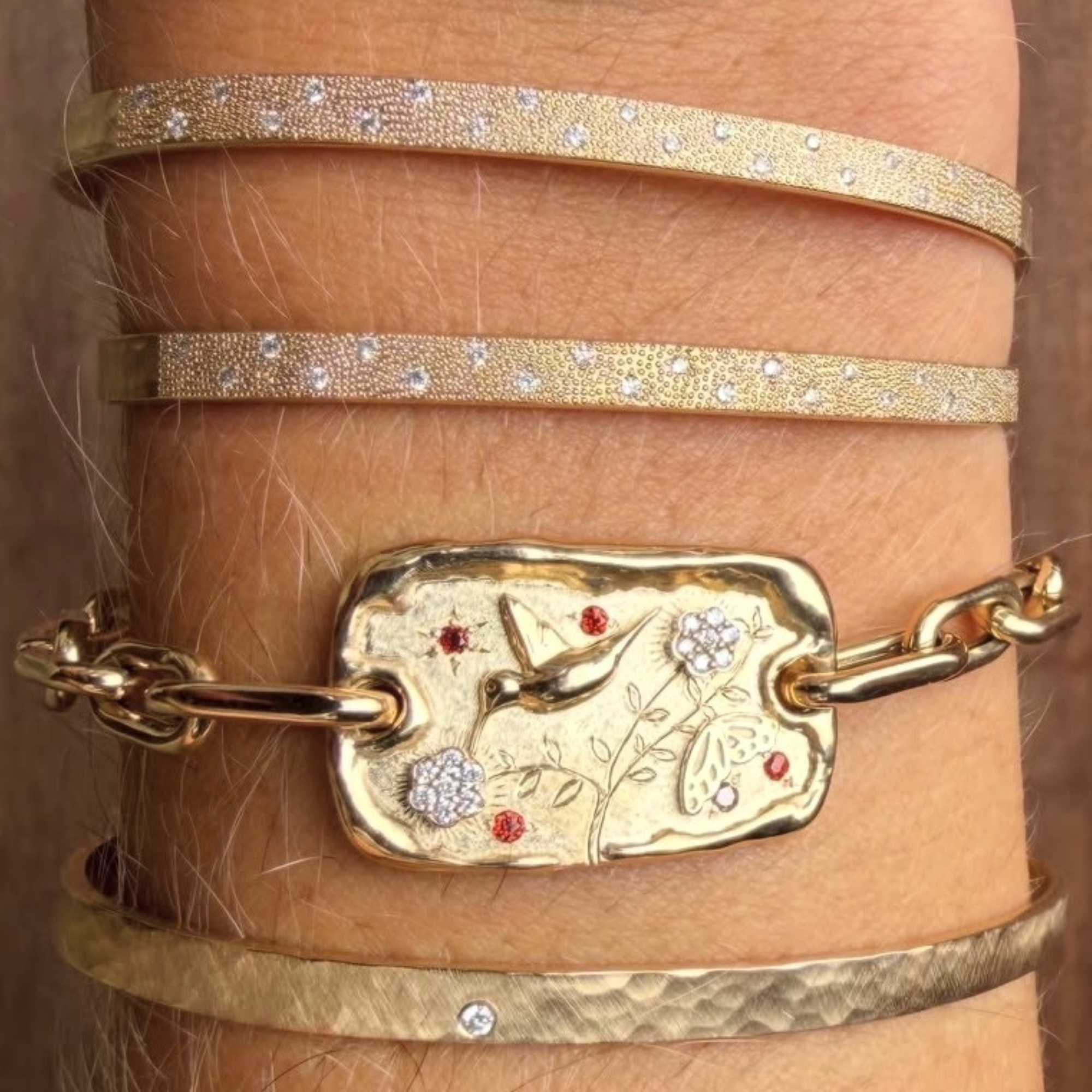 14k yellow gold charm chain bracelet with gold hummingbird and butterfly details and white diamond flowers and red garnet accent on a mica52 chain on wrist.