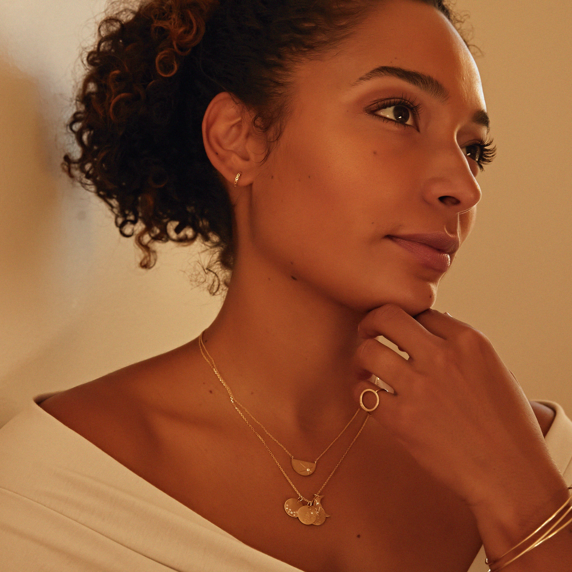 Woman wearing gold jewelry featuring chain lonk huggie hoop earrings and layered baby charm necklace with a warm-toned background