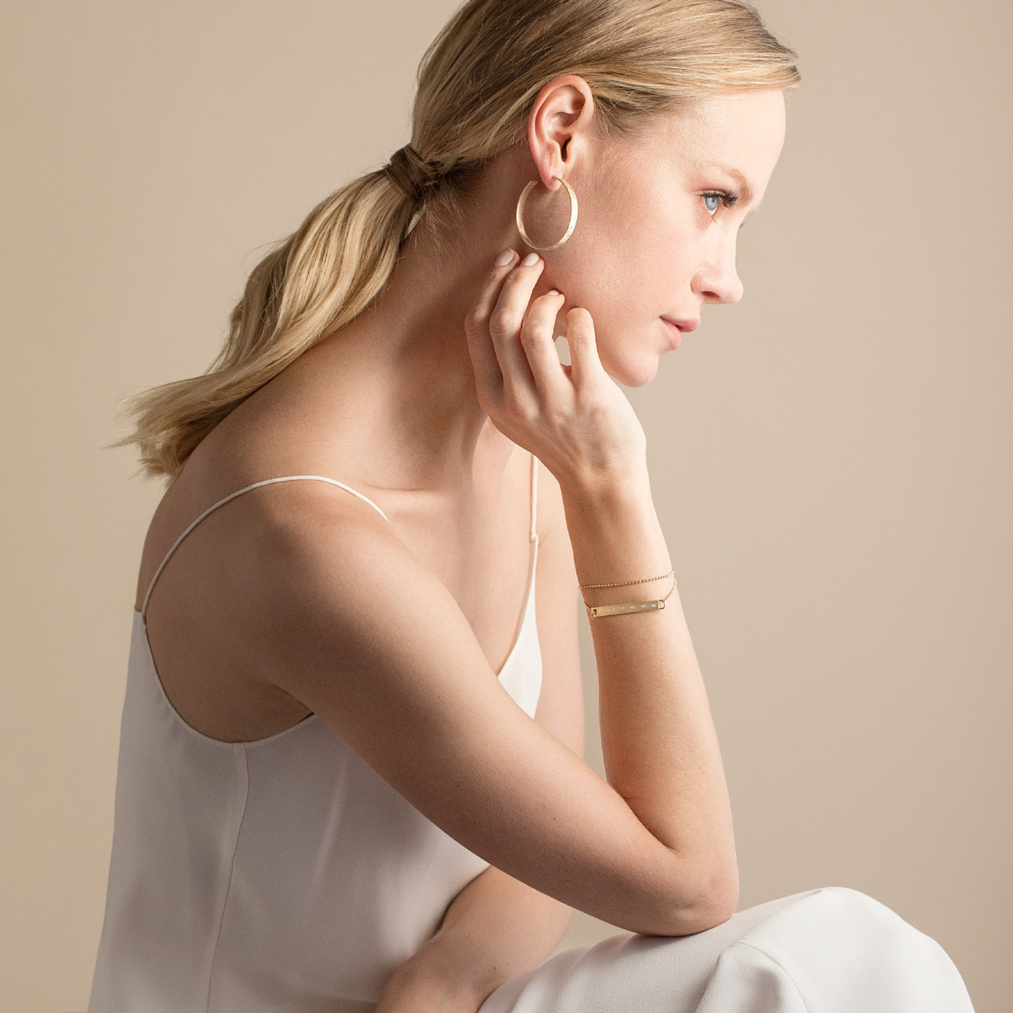 Woman wearing large yellow gold hoop earrings and double wrap bar chain bracelet white diamond accents against a beige background