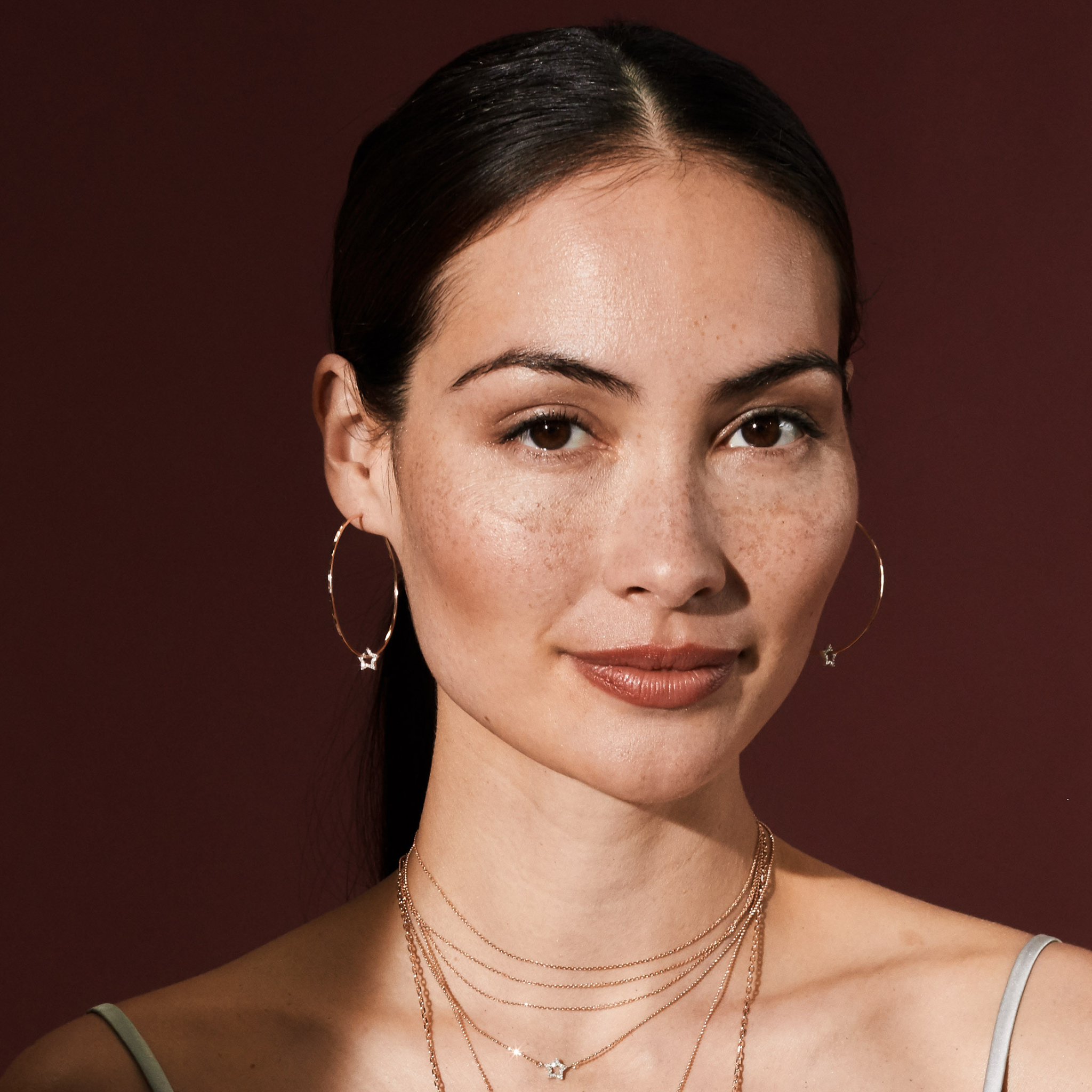 Woman wearing matching gold hoop earrings and necklaces with pave diamond star charms against a dark red background