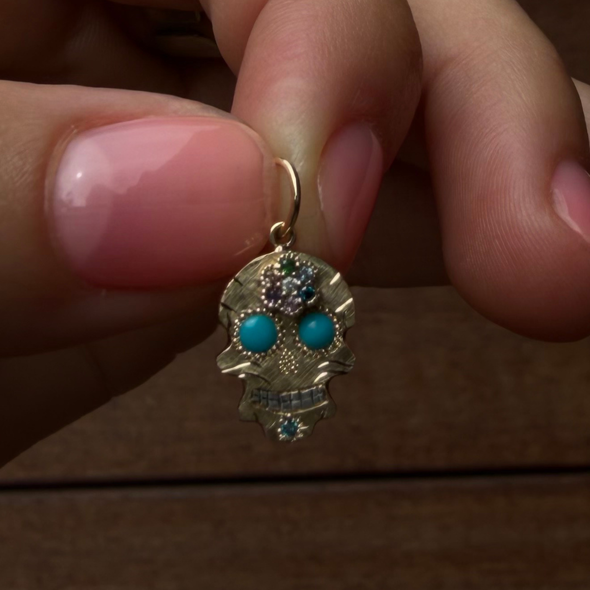 A close-up of a hand holding a small gold sugar skull pendant with turquoise eyes and sapphire flower details against a dark wooden background.