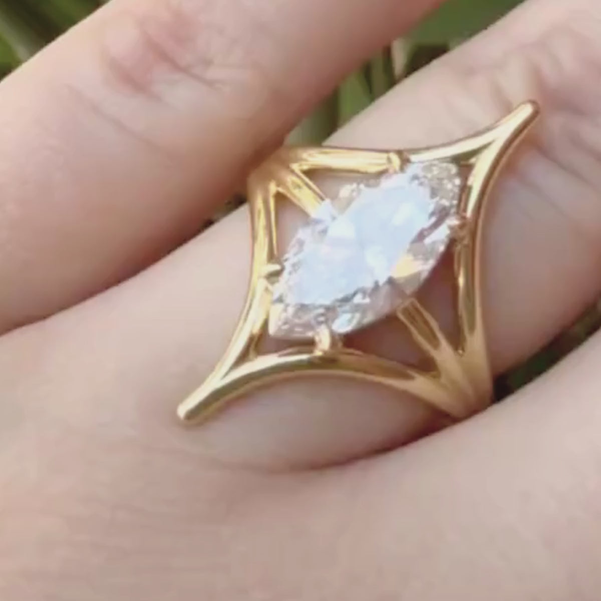 Candid video of hand close up wearing open cage marquise diamond solitaire engagement ring in yellow gold with blurred greenery background.