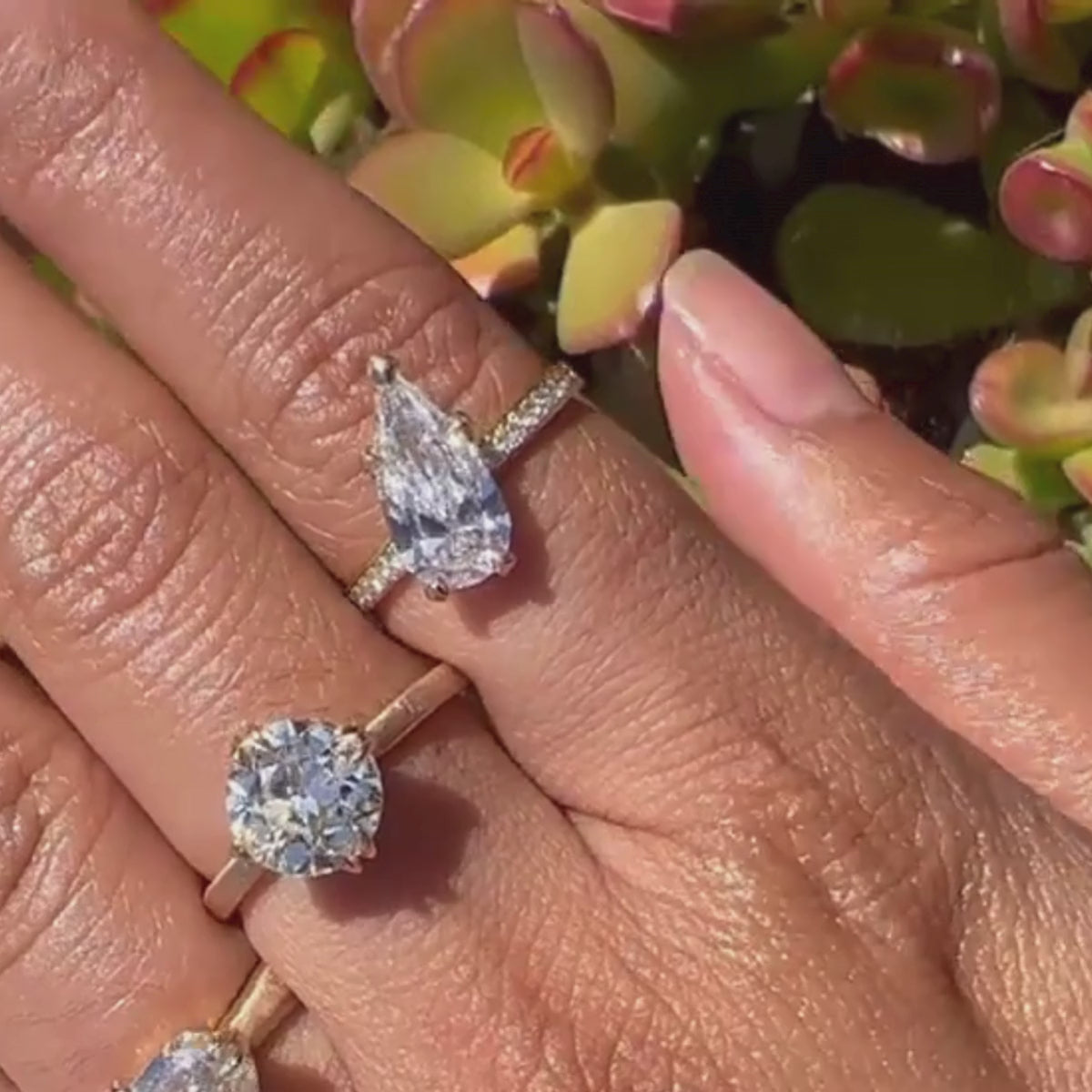 Candid video of elongated pear-cut-solitaire with tilted center stone and pave eternity band shown close-up on hand with succulents in the background.