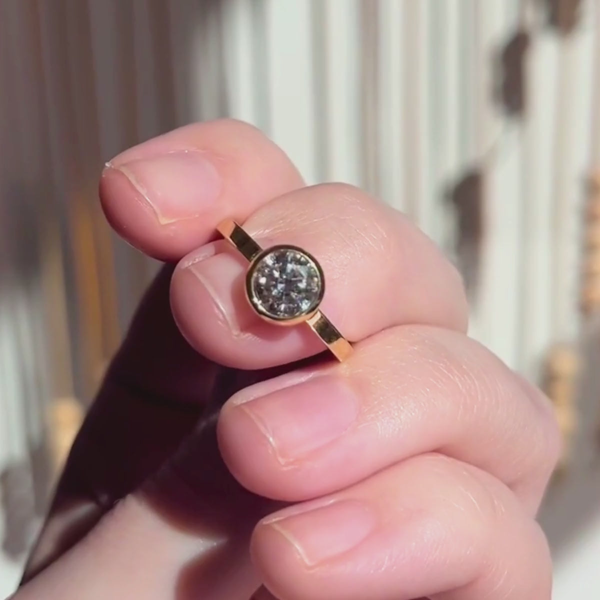 Candid video of hand holding round brilliant bezel solitaire engagement ring with square band blurred background.