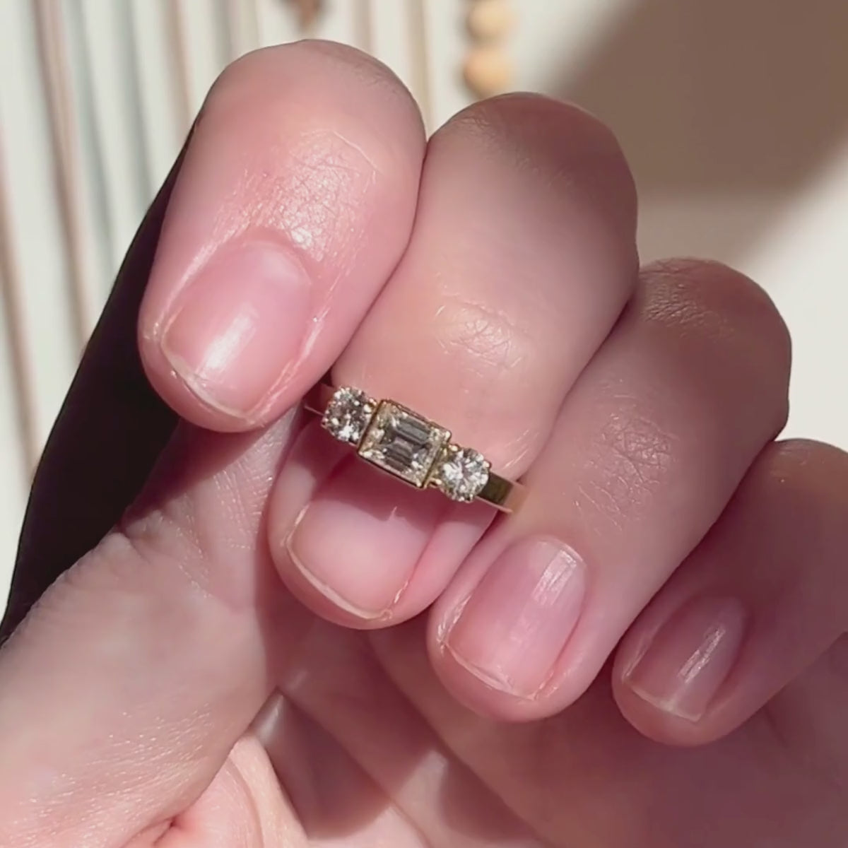 Candid video of three stone emeral-cut bezel set center stone and prong set round brilliant accent stones shown close-up on hand with blurred background.