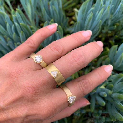 Hand wearing three 14k gold rings with diamonds wide cigar bands with brushed satin finish featuring single stone designs in pear, trillion and round shapes against a green plant background