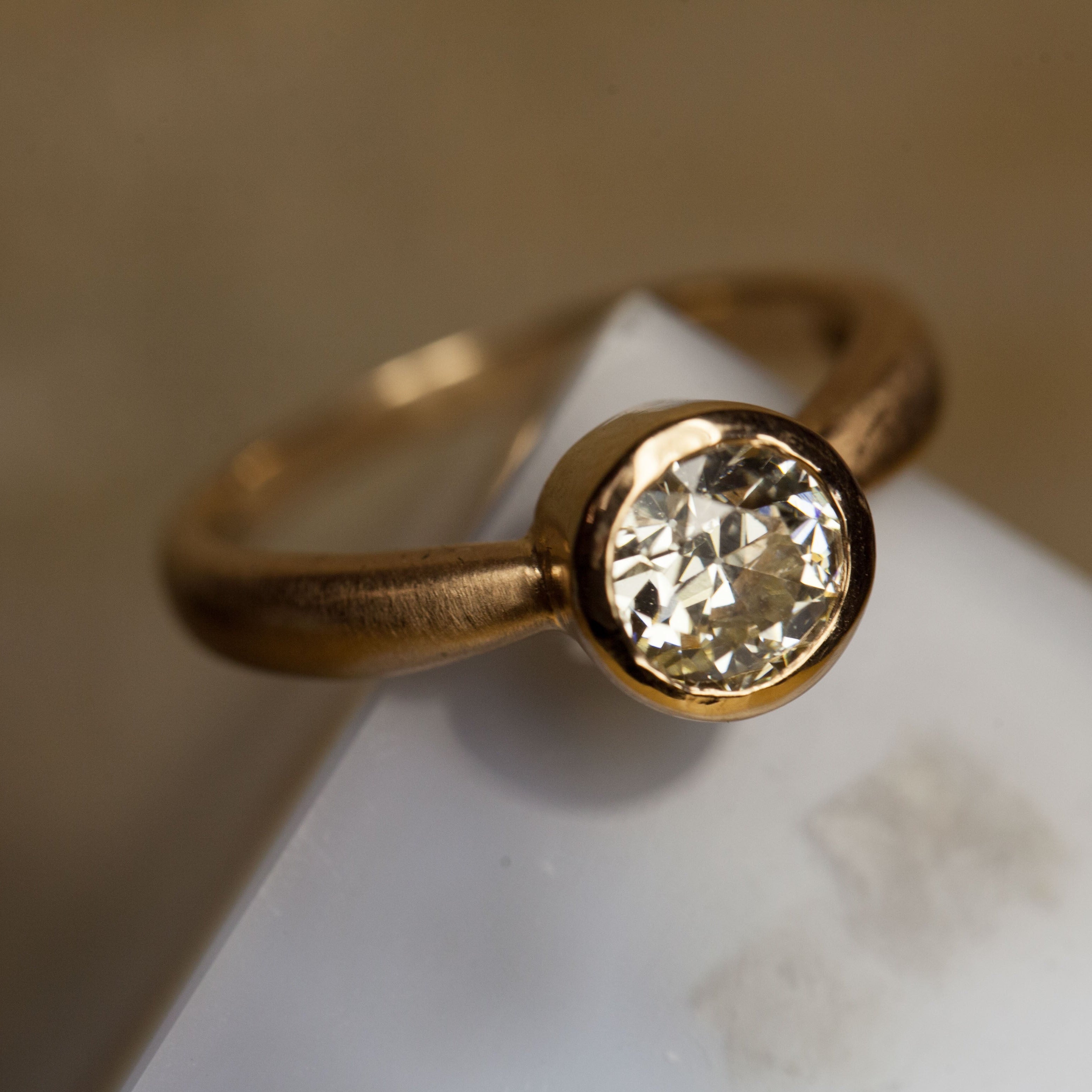 Yellow gold single stone solitaire round white diamond engagement ring with bezel setting brushed finish puffed band on white marble with blurred background