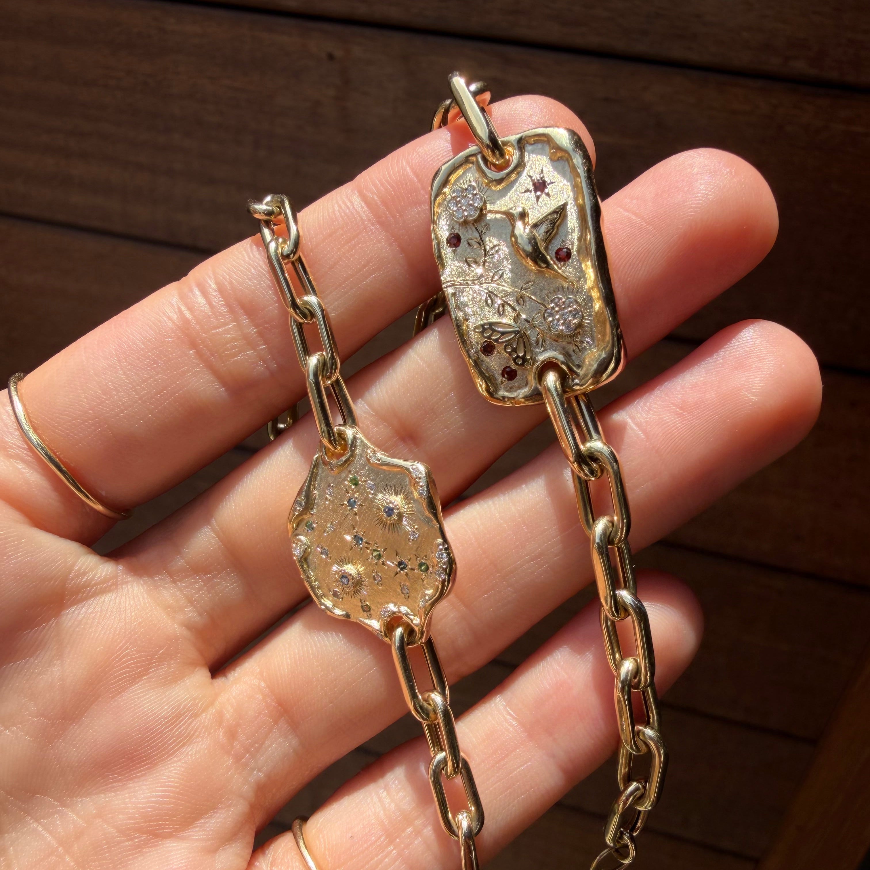 A close-up of a hand holding two gold-toned chain bracelets with irregular shield-shaped charms featuring textured star patterns and small red gemstone accents, set against a dark wooden background.