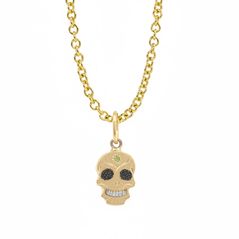 A Teeny Tiny collection gold charm in the shape of a sugar skull with a round apple green diamond in the center, displayed on a gold chain against a white background.