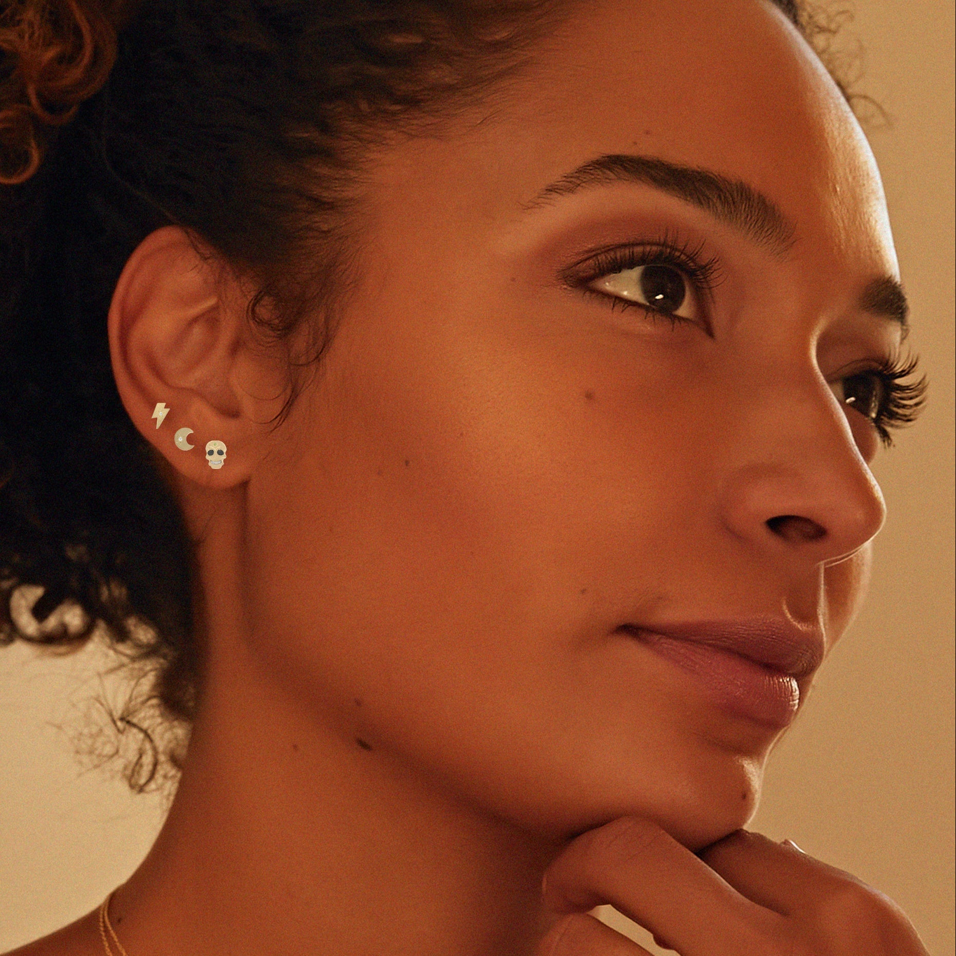 Close-up of a woman wearing teeny tiny collection of 14k gold earrings warm-toned background.