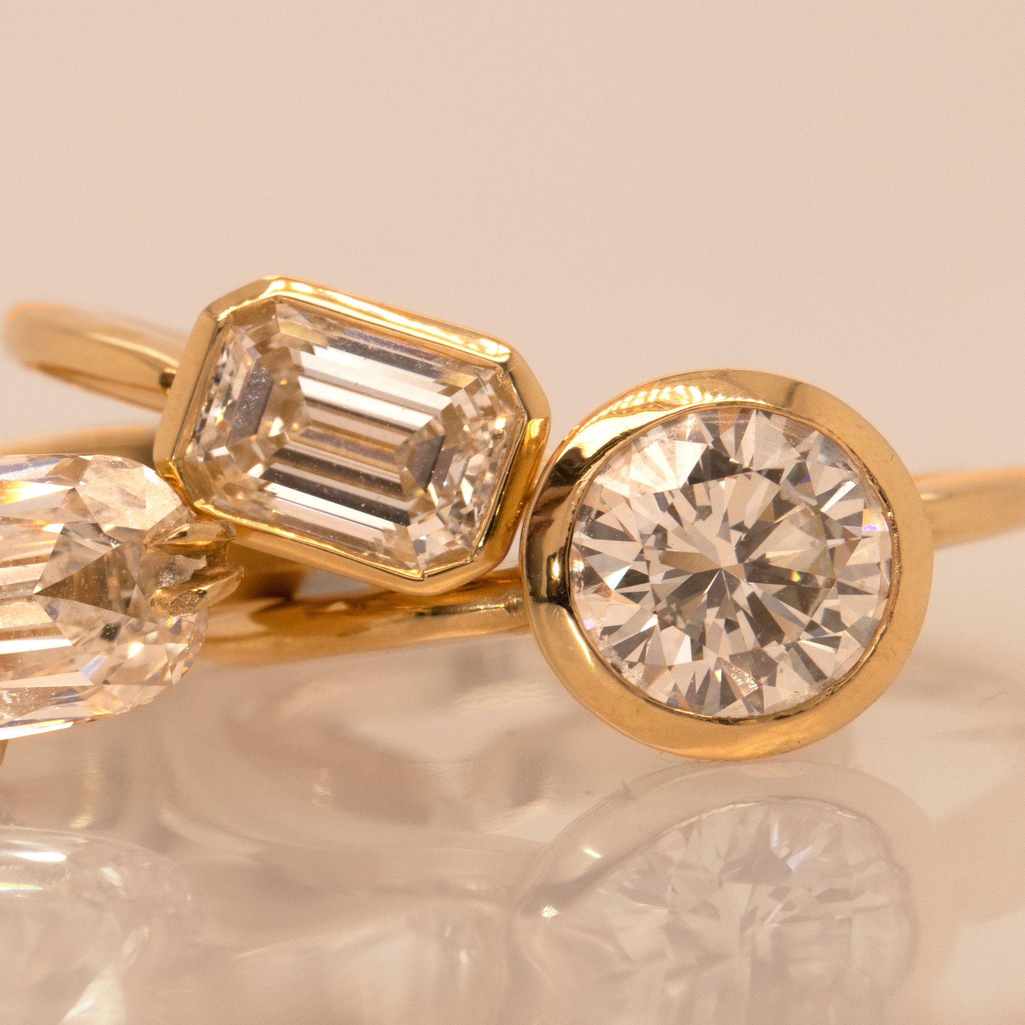 Round brilliant and emerals diamond solitaire bezel engagement rings in yellow gold reflective blush background