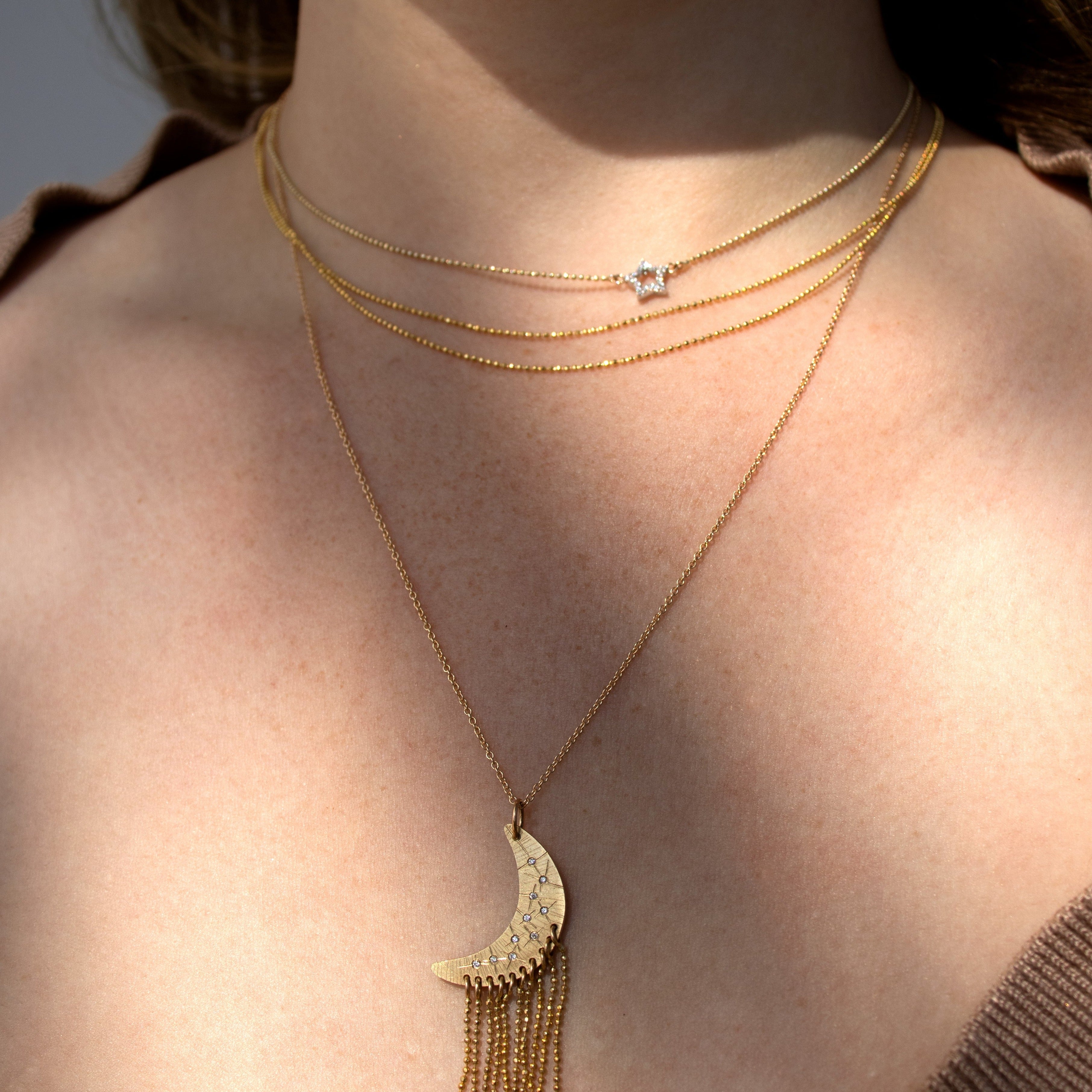 Model wearing layered necklaces. Dainty three strand diamond ball chain ckoker with pave set white diamond star. Paired with longer necklae featuring a diamond and yellow gold oon pendant with tassles of diamond ball chain.