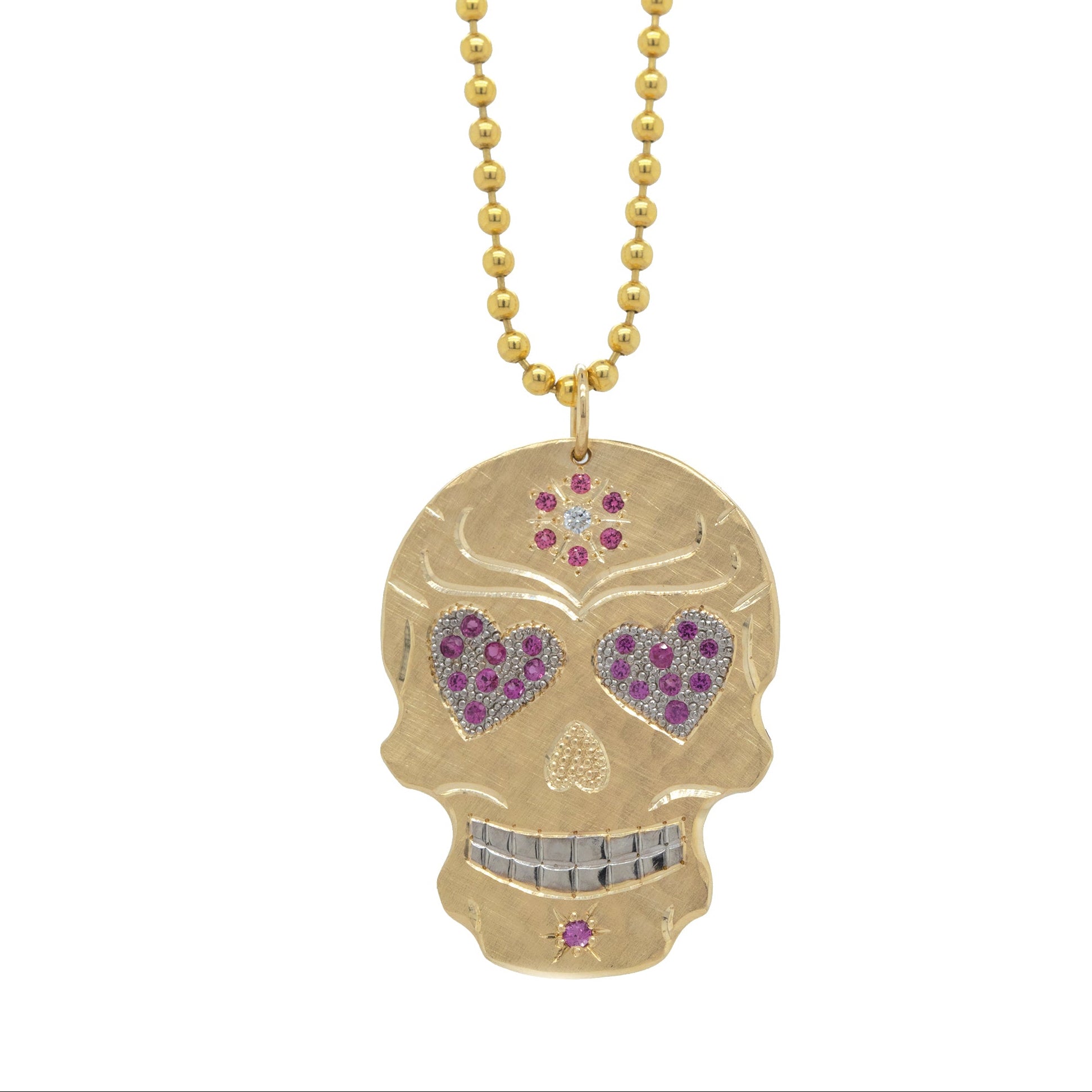 A gold-colored sugar skull pendant with heart-shaped eyes adorned with pink sapphires and spinel, suspended from a gold ball chain.