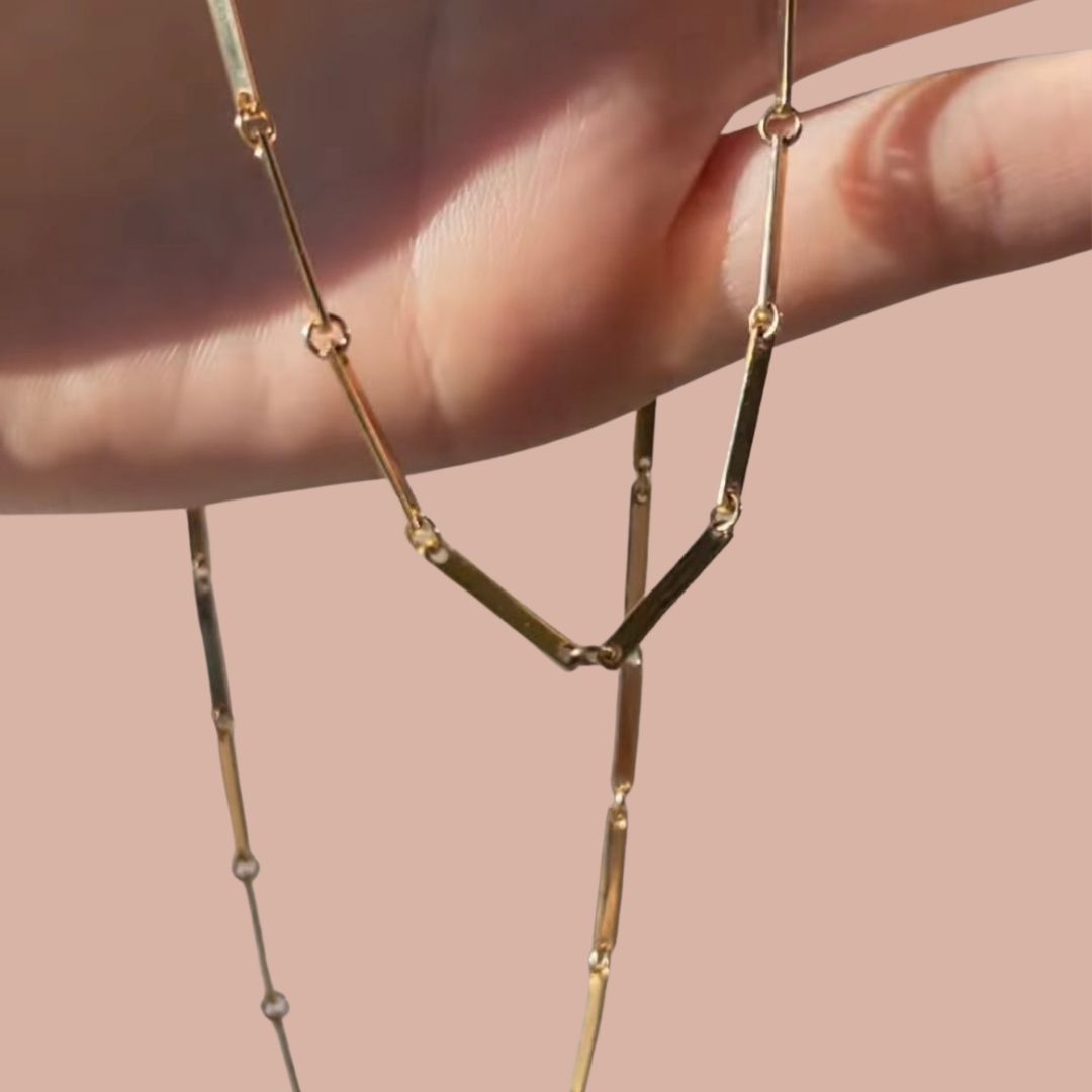 Hand holding long bar link chain necklace in yellow gold, pink background.